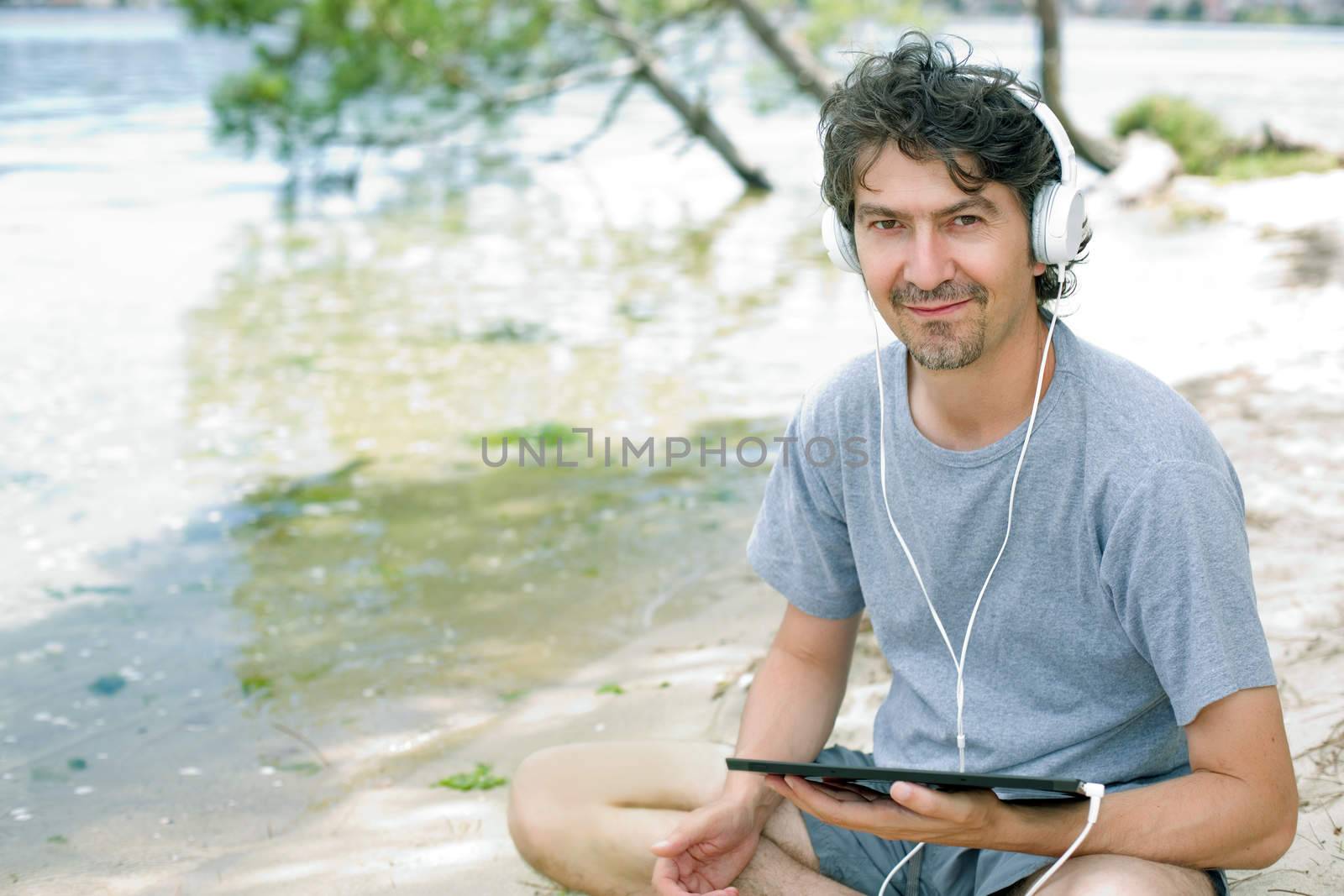 young man holding a tablet with headphones, at the beach