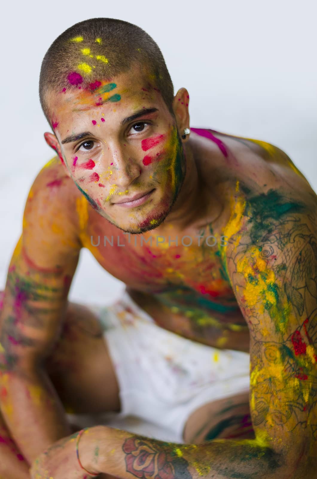 Attractive young man shirtless, skin painted all over with bright Holi colors, looking at camera, sitting, on white background