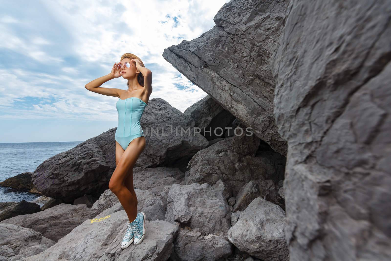 Beautiful young teen girl in bodysuit and hat posing on the stones in the sea fashion shoot at shore