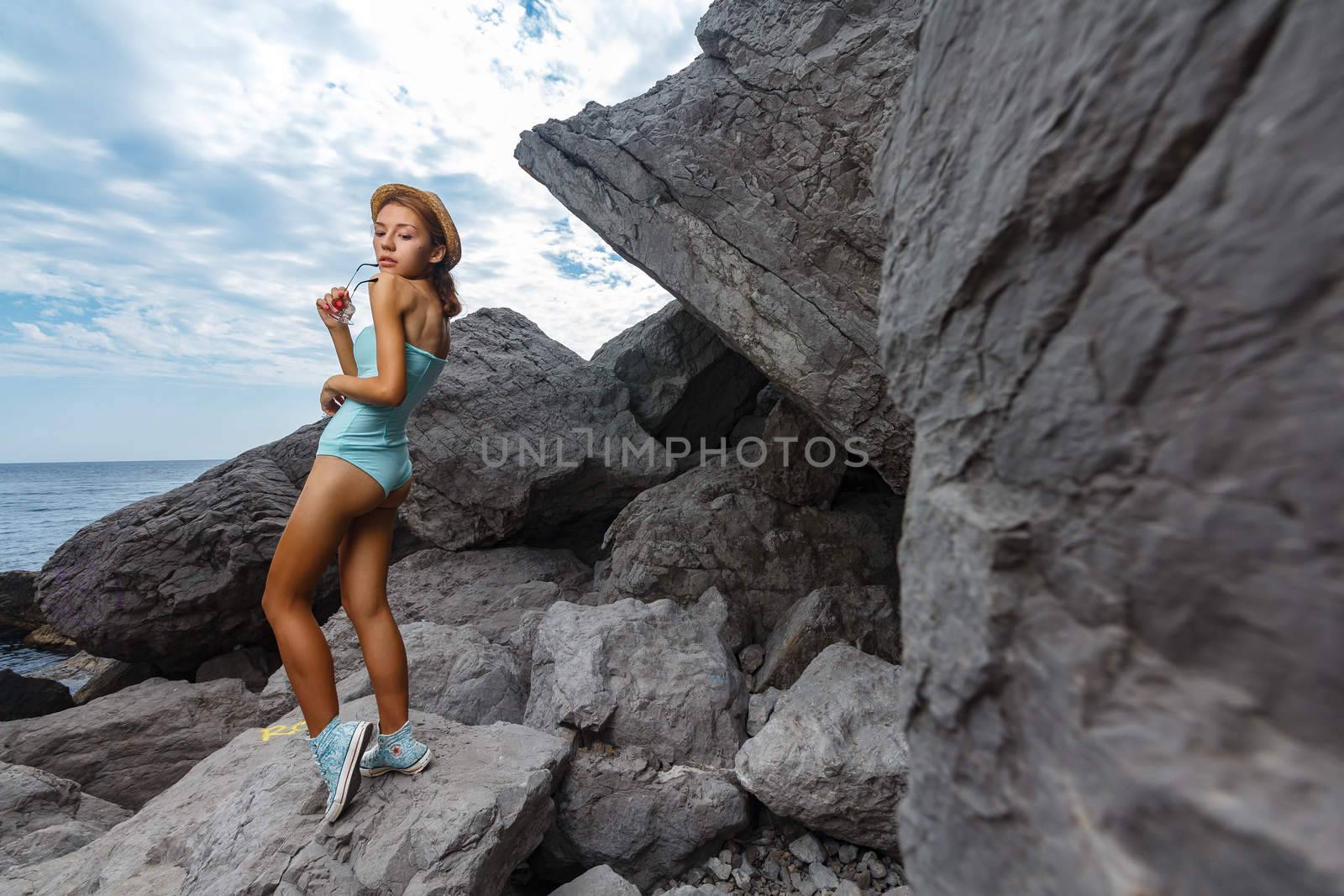 Beautiful young teen girl in bodysuit and hat posing on the stones in the sea fashion shoot at shore