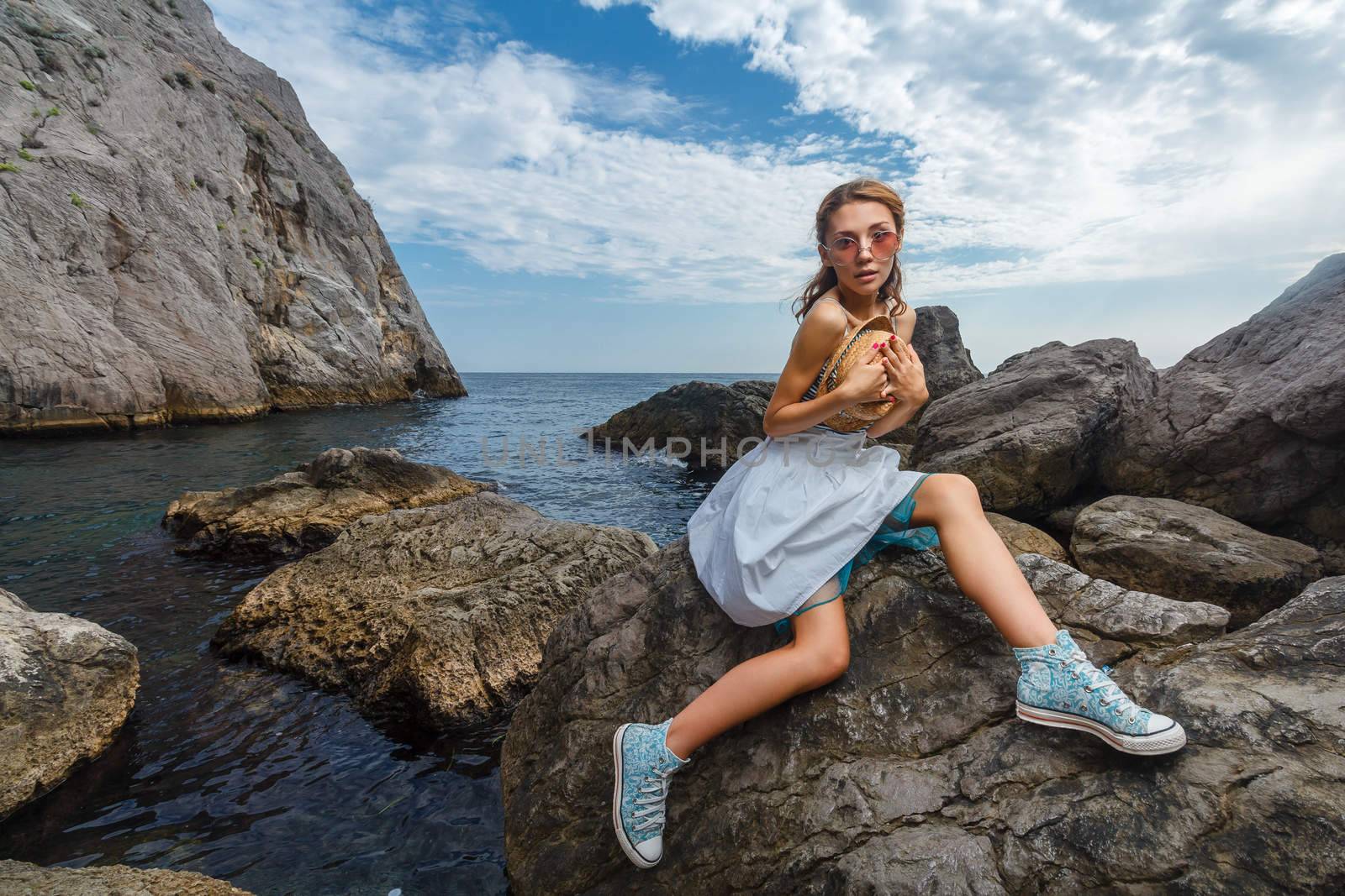 Beautiful young teen girl in dress and hat posing on the stones in the sea fashion shoot at shore
