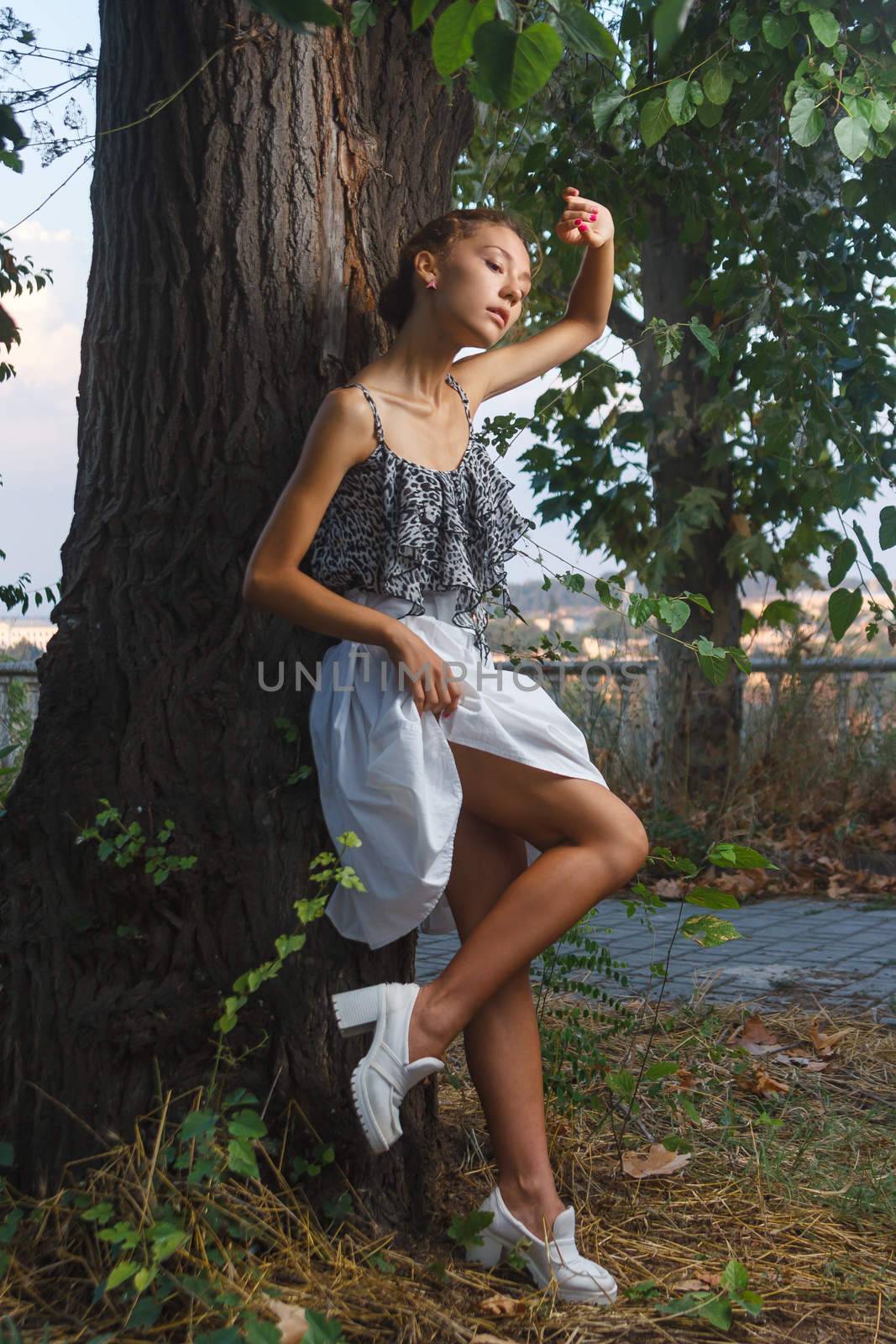 Fashion portrait shoot of a beautiful teen girl posing near tree during city fashion photoshoot with greenery at background