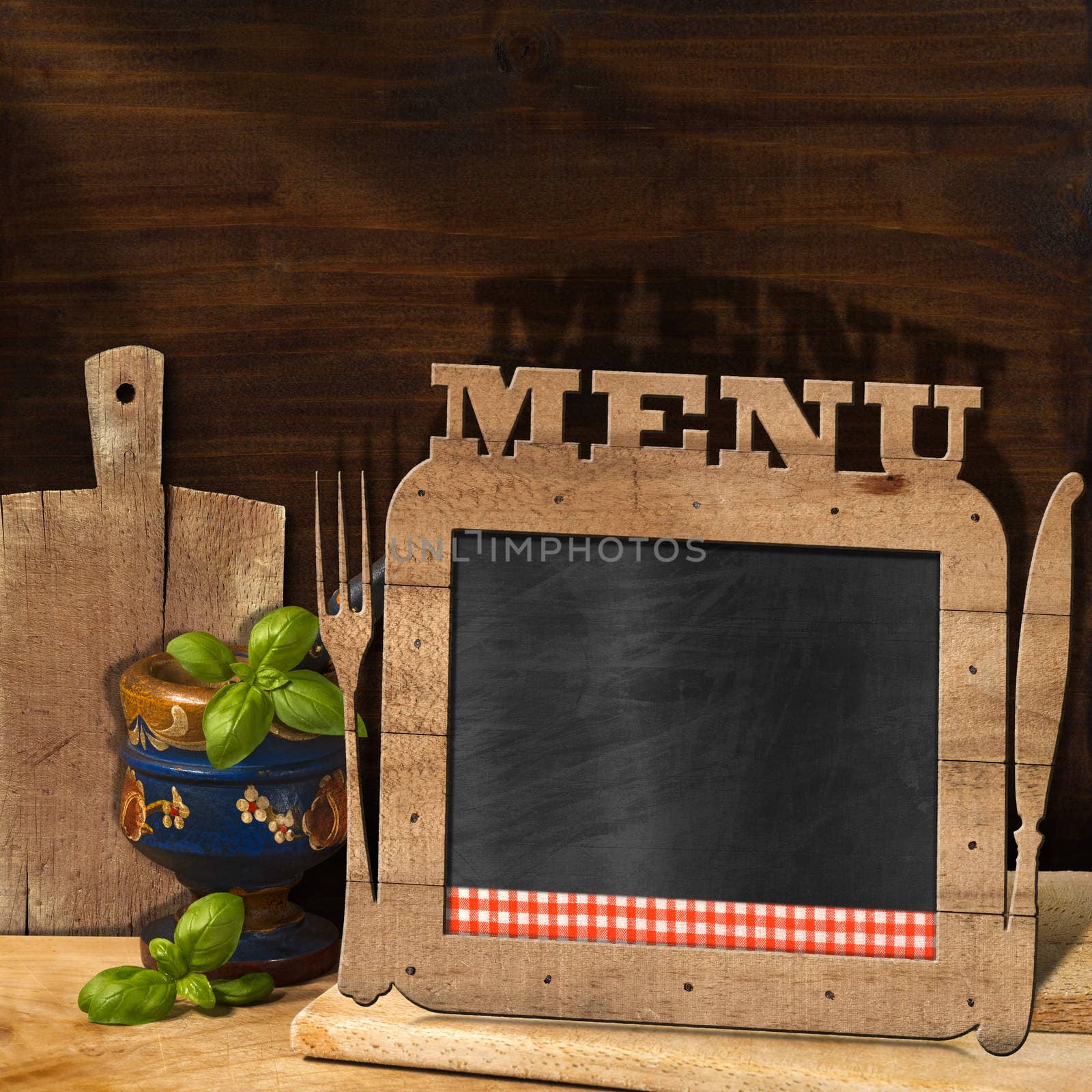 Empty blackboard with wooden frame, text Menu, fork and knife in the kitchen with mortar and pestle with basil leaves and cutting boards. Template for food menu