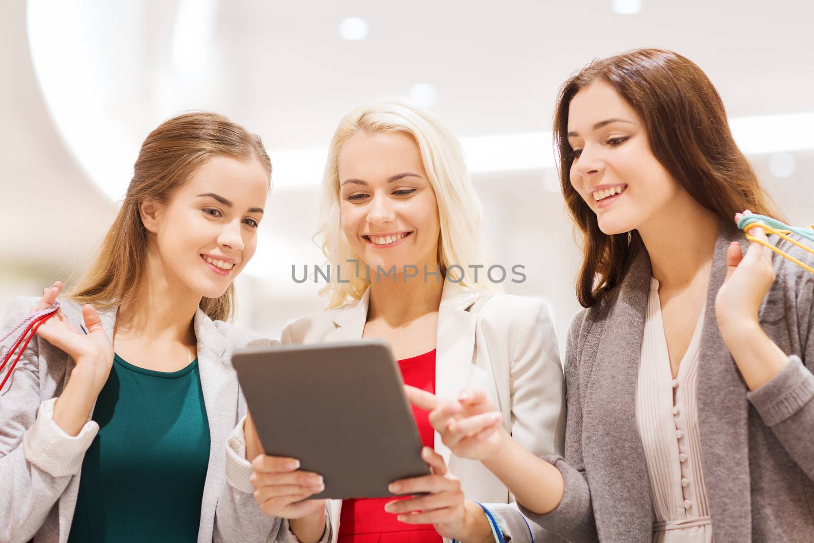 sale, consumerism, technology and people concept - happy young women with tablet pc and shopping bags in mall