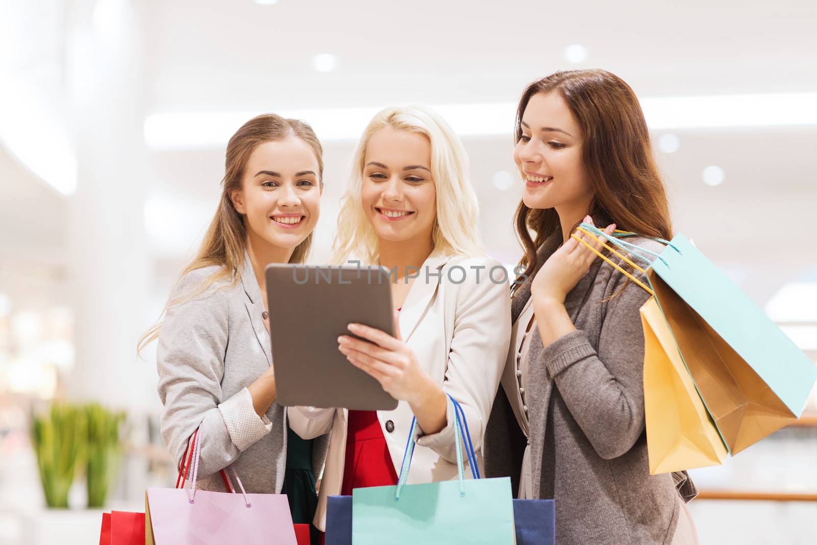 sale, consumerism, technology and people concept - happy young women with tablet pc and shopping bags in mall