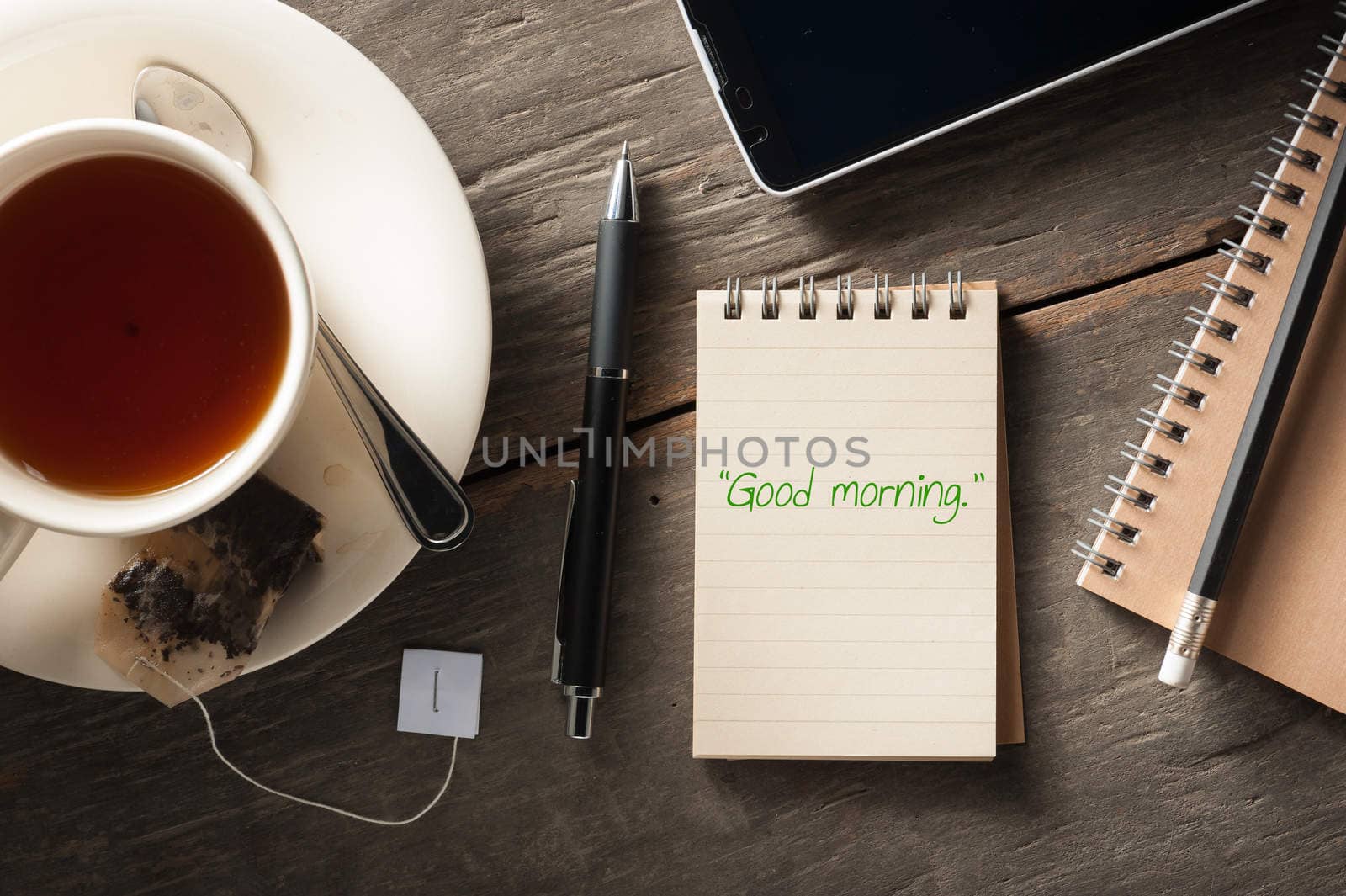 "Good morning" is written on small notepad with a cup of tea, pen, pencil and cellphne on rustic wood background in morning time with low key scene