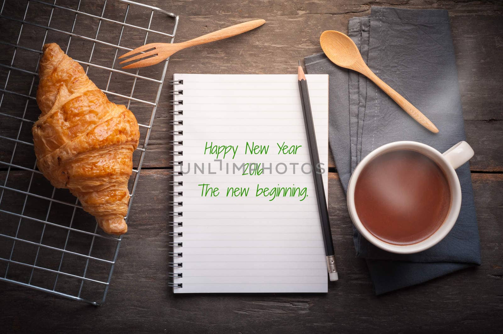 "Happy new year 2016 The new begining" is written on notebook with Butter croissant, a cup of hot chocolate on wood table in morning time