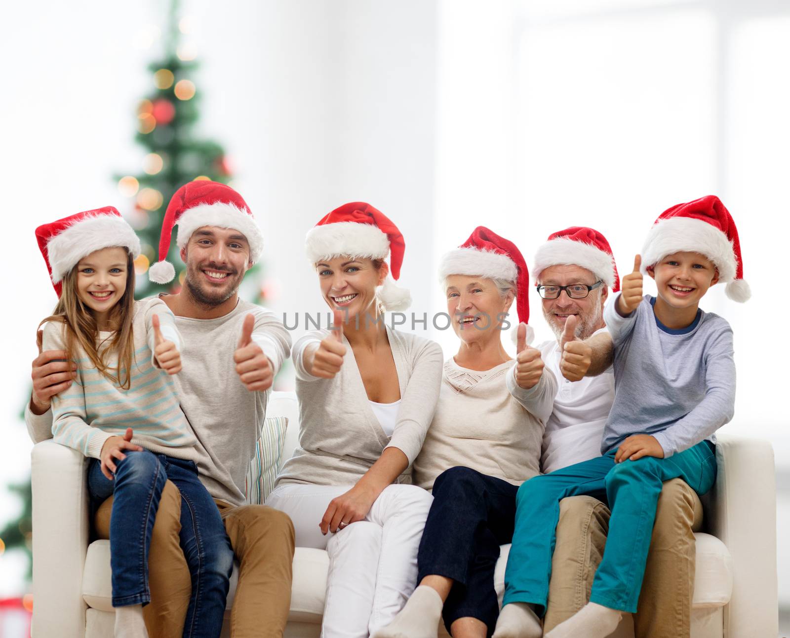 family, home, generation, holidays and people concept - happy family in santa helper hats sitting on couch and showing thumbs up gesture over living room with christmas tree background