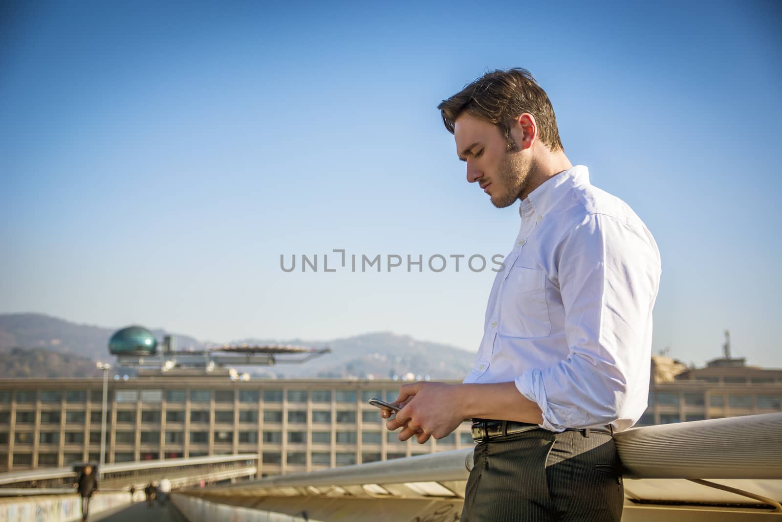 Handsome trendy man wearing white shirt standing and looking down at a cell phone that he is holding, outdoor in city setting