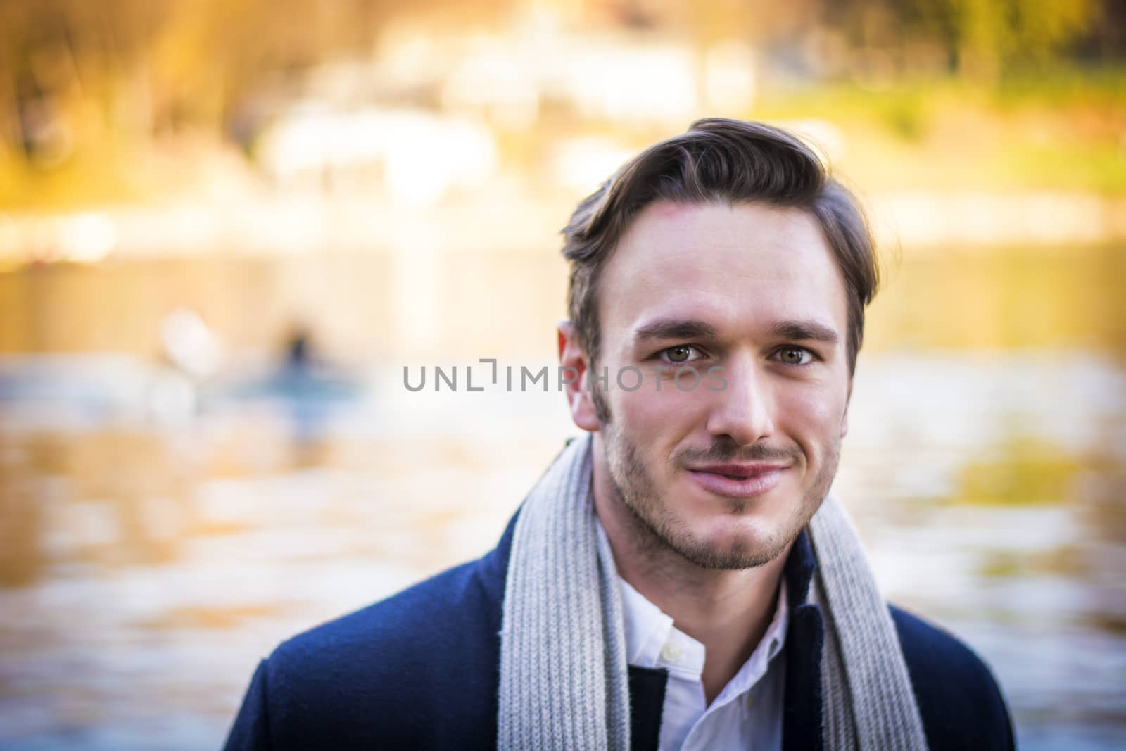 Handsome young man outdoor in winter fashion, wearing black coat and woolen scarf next to lake or river, smiling to camera