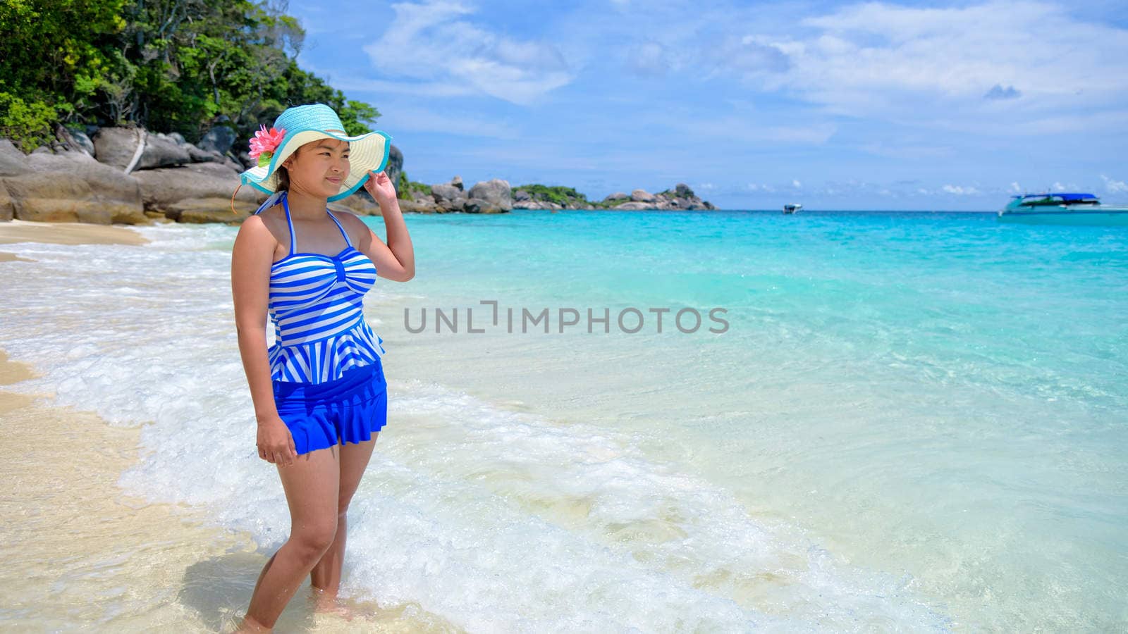 Beautiful woman in a blue striped swimsuit and hat standing poses on beach of sea under a summer sky at Koh Miang Island in Mu Ko Similan National Park, Phang Nga Province, Thailand, 16:9 widescreen