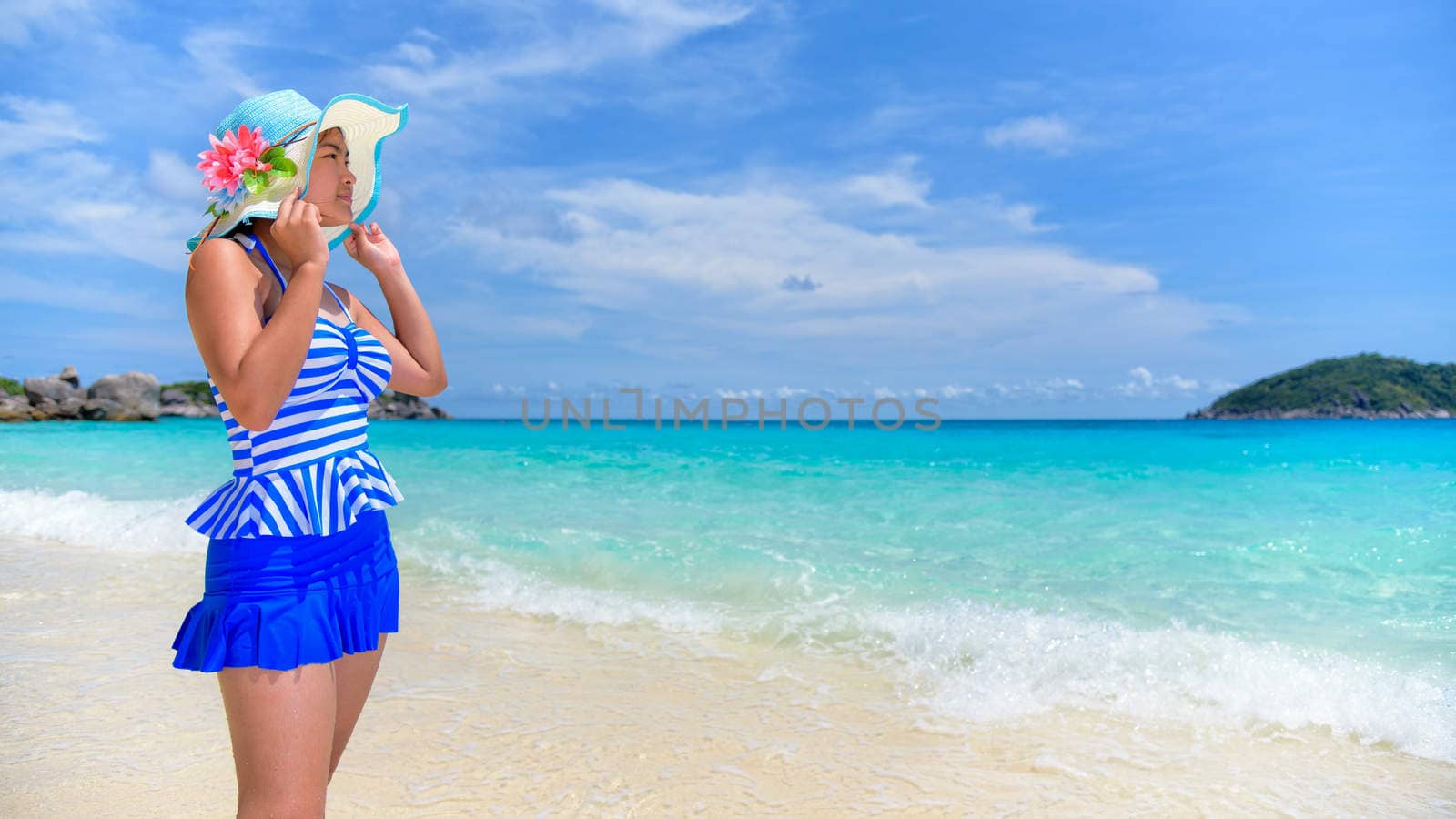 Beautiful woman in a blue striped swimsuit and hat standing poses on beach of sea under a summer sky at Koh Miang Island in Mu Ko Similan National Park, Phang Nga Province, Thailand, 16:9 widescreen