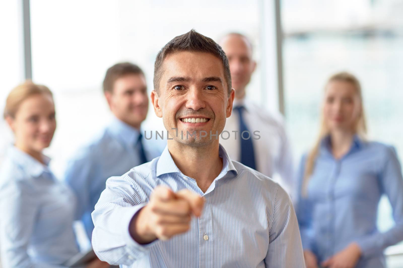 business, people, gesture and teamwork concept - smiling businessman pointing finger to you with group of businesspeople meeting in office