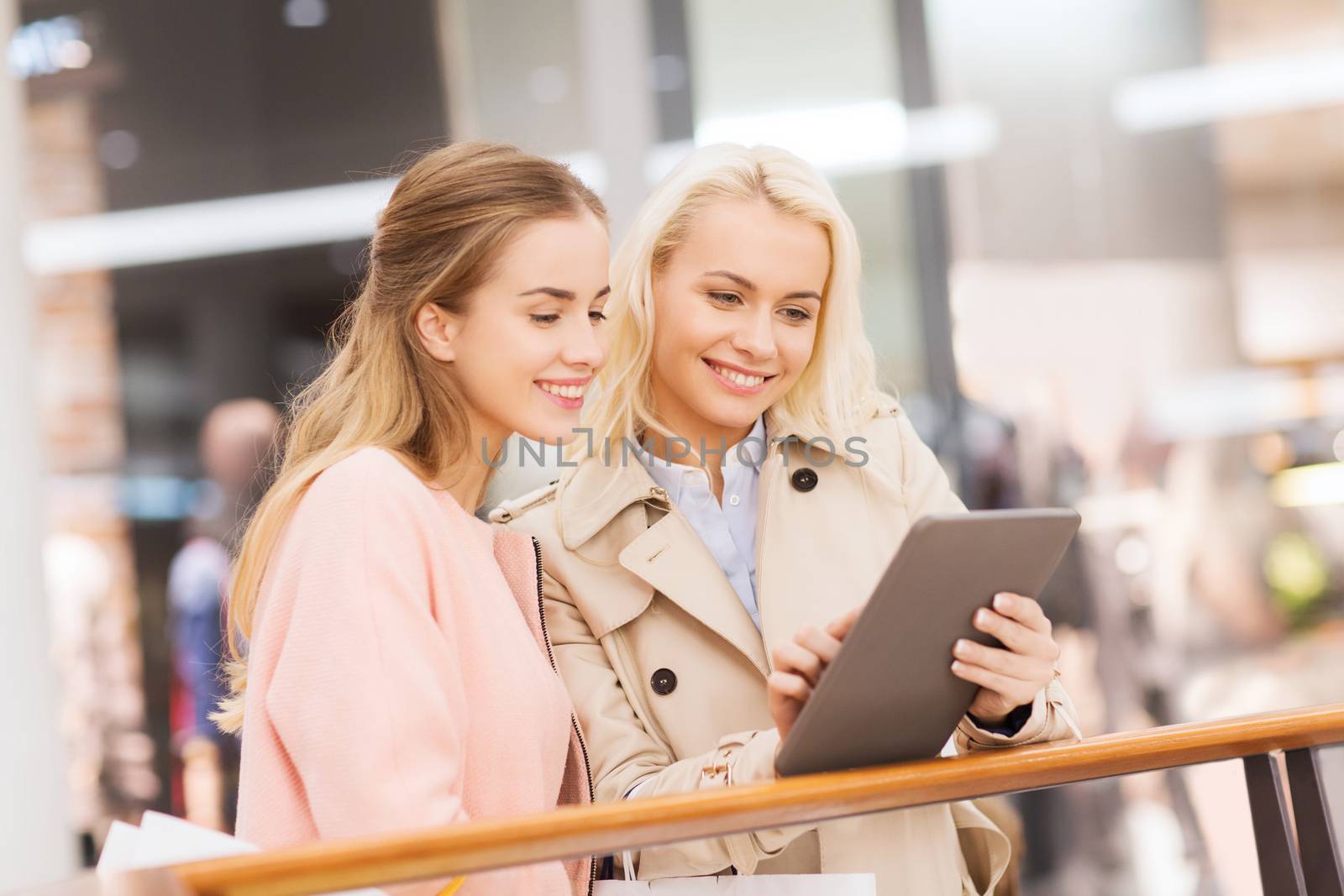 sale, consumerism, technology and people concept - happy young women with tablet pc and shopping bags in mall