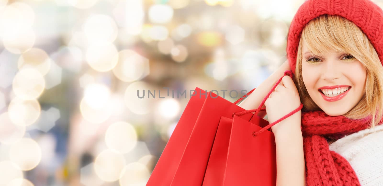 happiness, winter holidays, christmas and people concept - smiling young woman in hat and scarf with red shopping bags over lights background