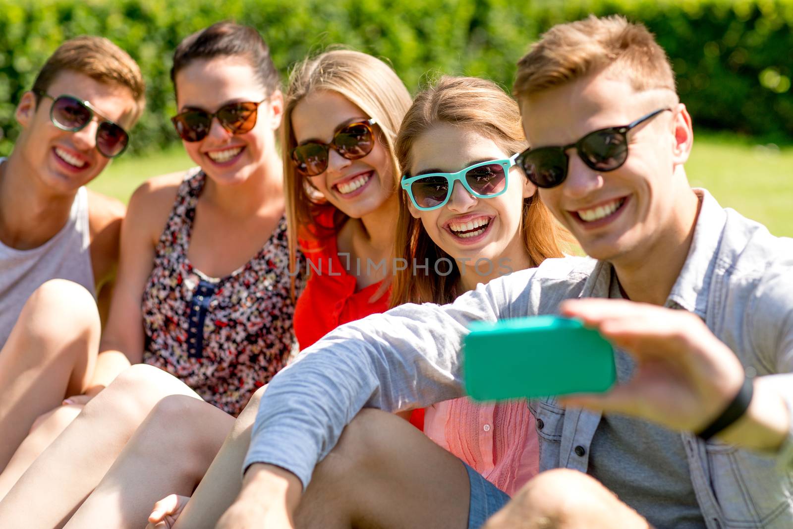 smiling friends with smartphone sitting on grass by dolgachov