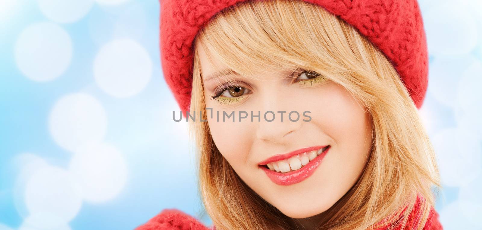 happiness, winter holidays, christmas and people concept - close up of smiling young woman in red hat and scarf over blue lights background