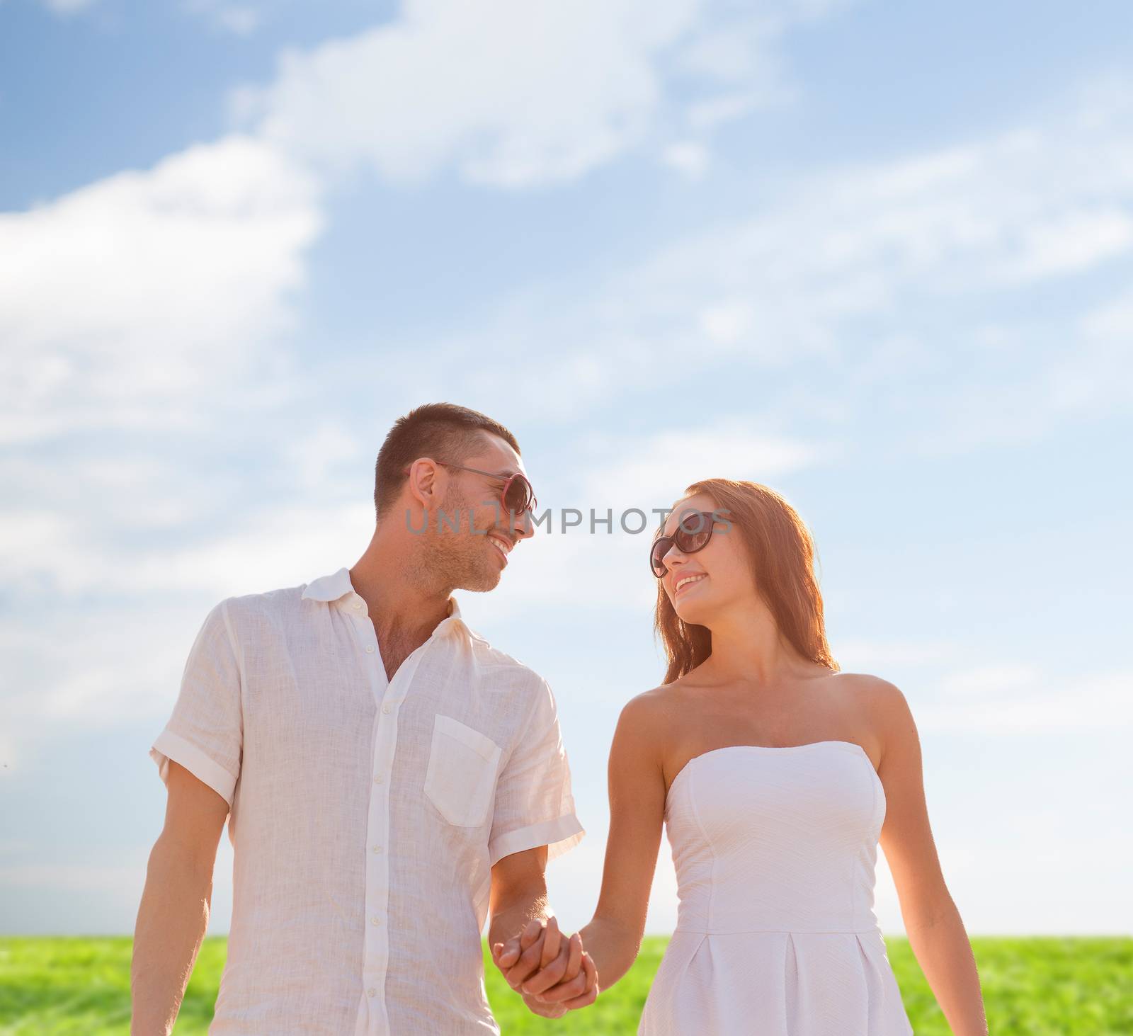 love, people, summer and relations concept - smiling couple wearing sunglasses walking outdoors over blue sky and grass background