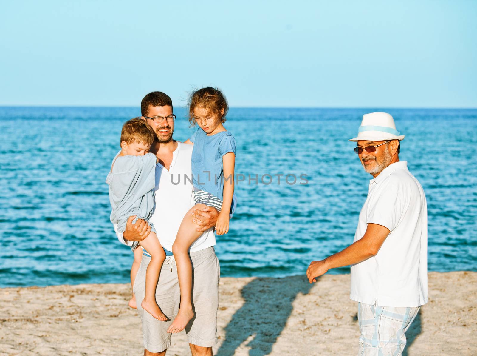 A father is lifting brother and sister kids on the seashore while grandfather is approaching with a smile.