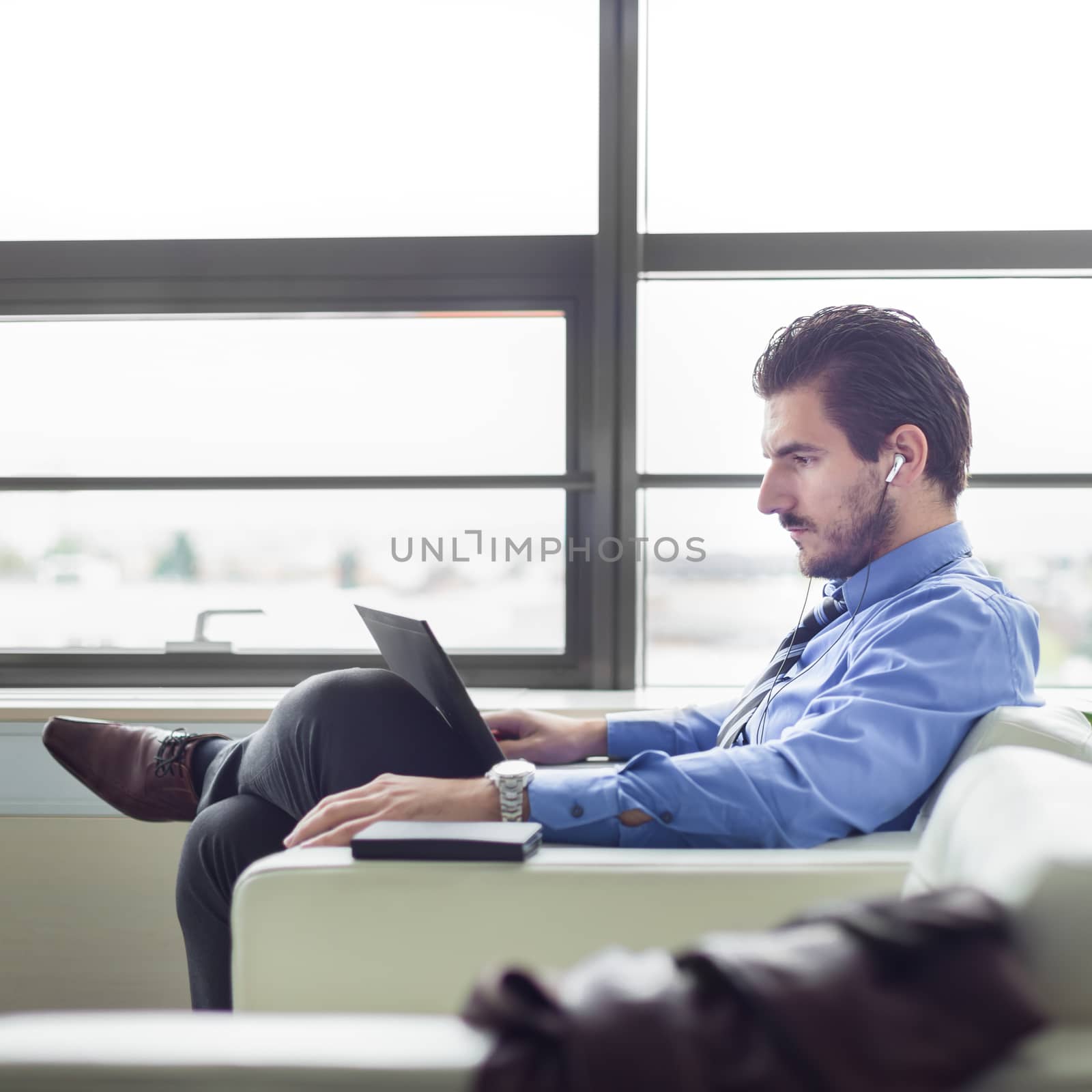 Portrait of successful young businessman in bright modern office focused on work on his laptop computer wearing headphones. Side view. Business and entrepreneurship concept.