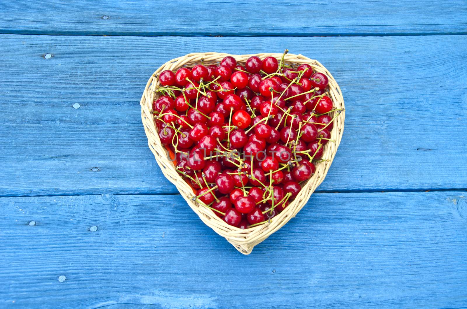 Heart shaped wicker basket full of cherries on blue old garden table wooden background