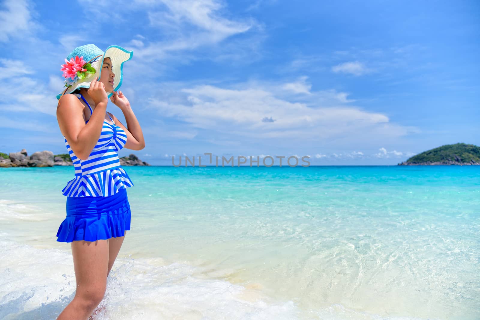 Beautiful woman in a blue striped swimsuit and hat standing poses on beach of sea under a summer sky at Koh Miang Island in Mu Ko Similan National Park, Phang Nga Province, Thailand