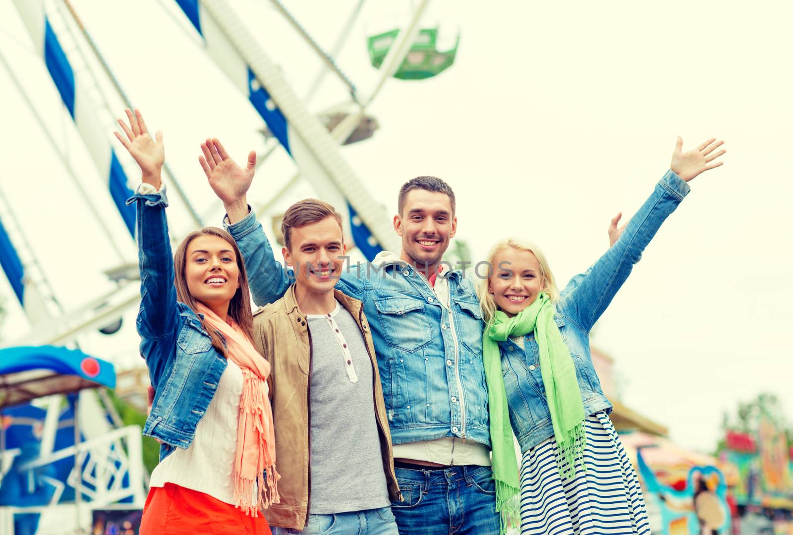 leisure, amusement park and friendship concept - group of smiling friends waving hands with ferris wheel on the back