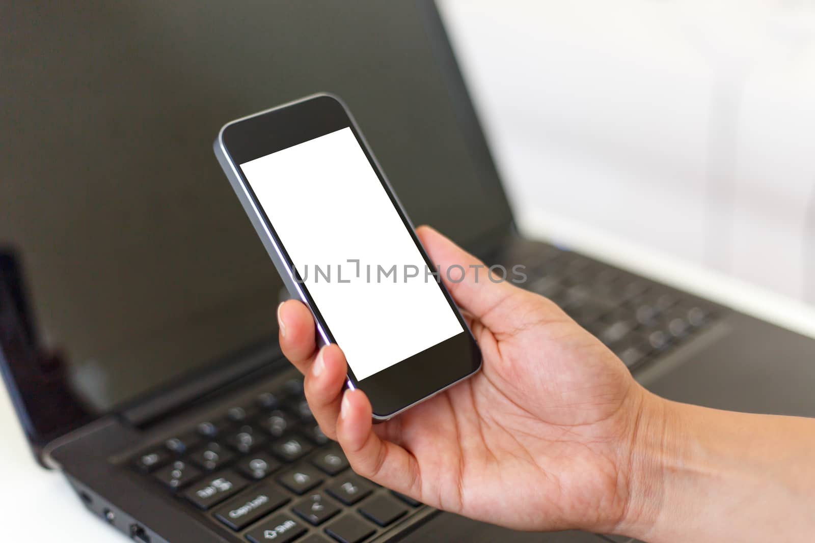 Close up detailed view of a smart phone holding on hand, with white blank copy space screen, on laptop background.