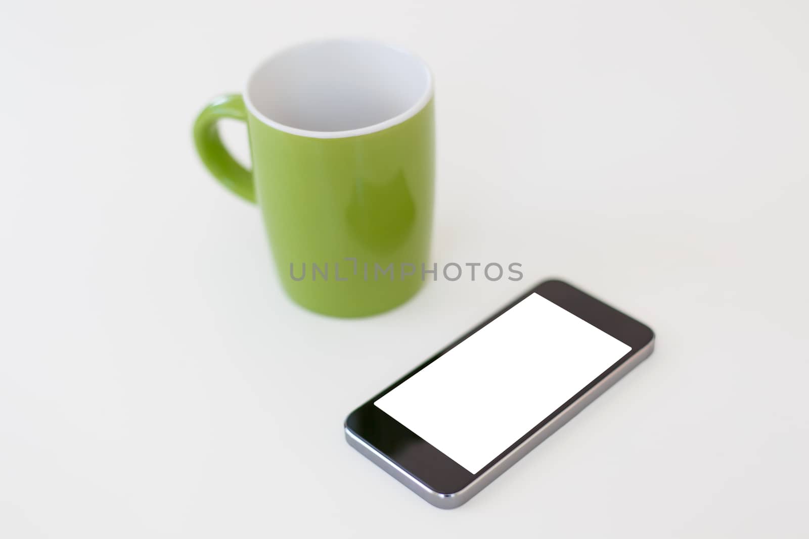 Close up detailed view of a smart phone, with white blank copy space screen, near a green mug, on white background.