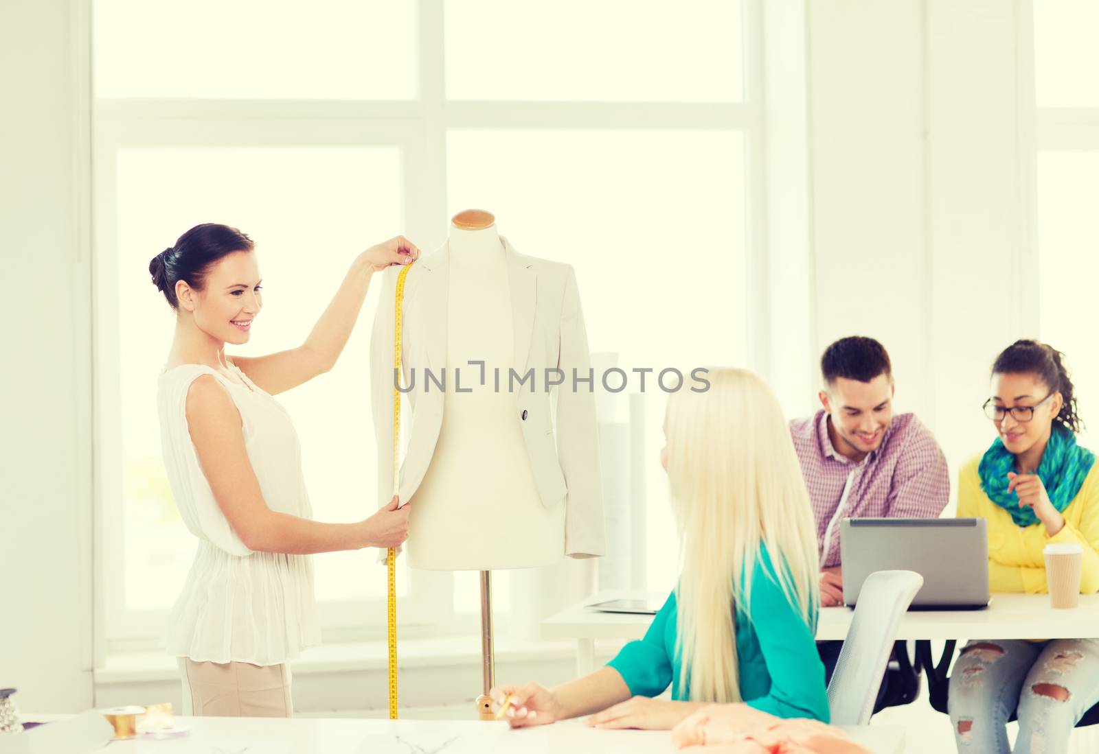 startup, education, fashion and office concept - smiling designers drawing sketches and measuring jacket on mannequin in office