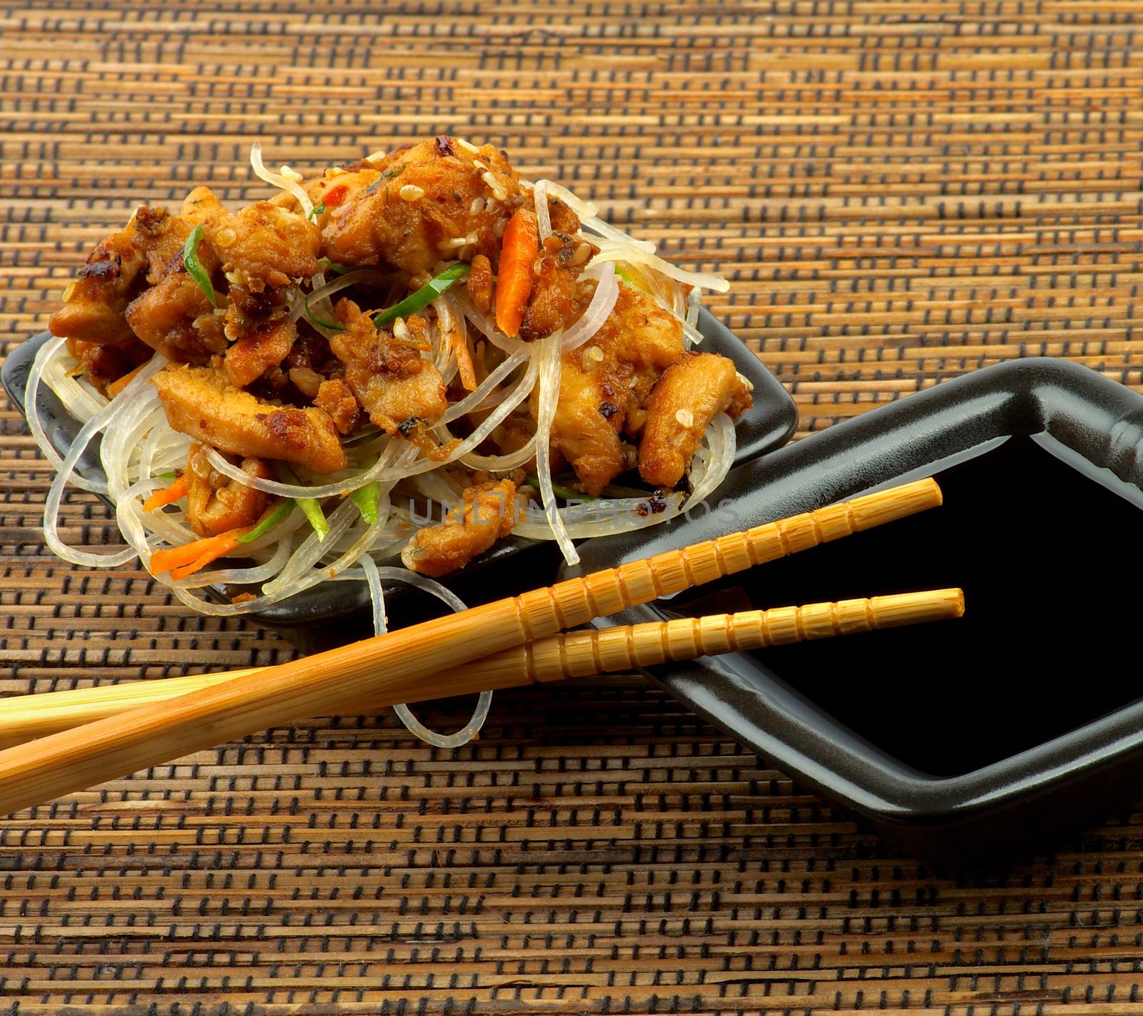 Delicious Chicken Teriyaki with Chinese Glass Noodles and Soy Sauce in Black Bowls with Chopsticks closeup on Strawmat background