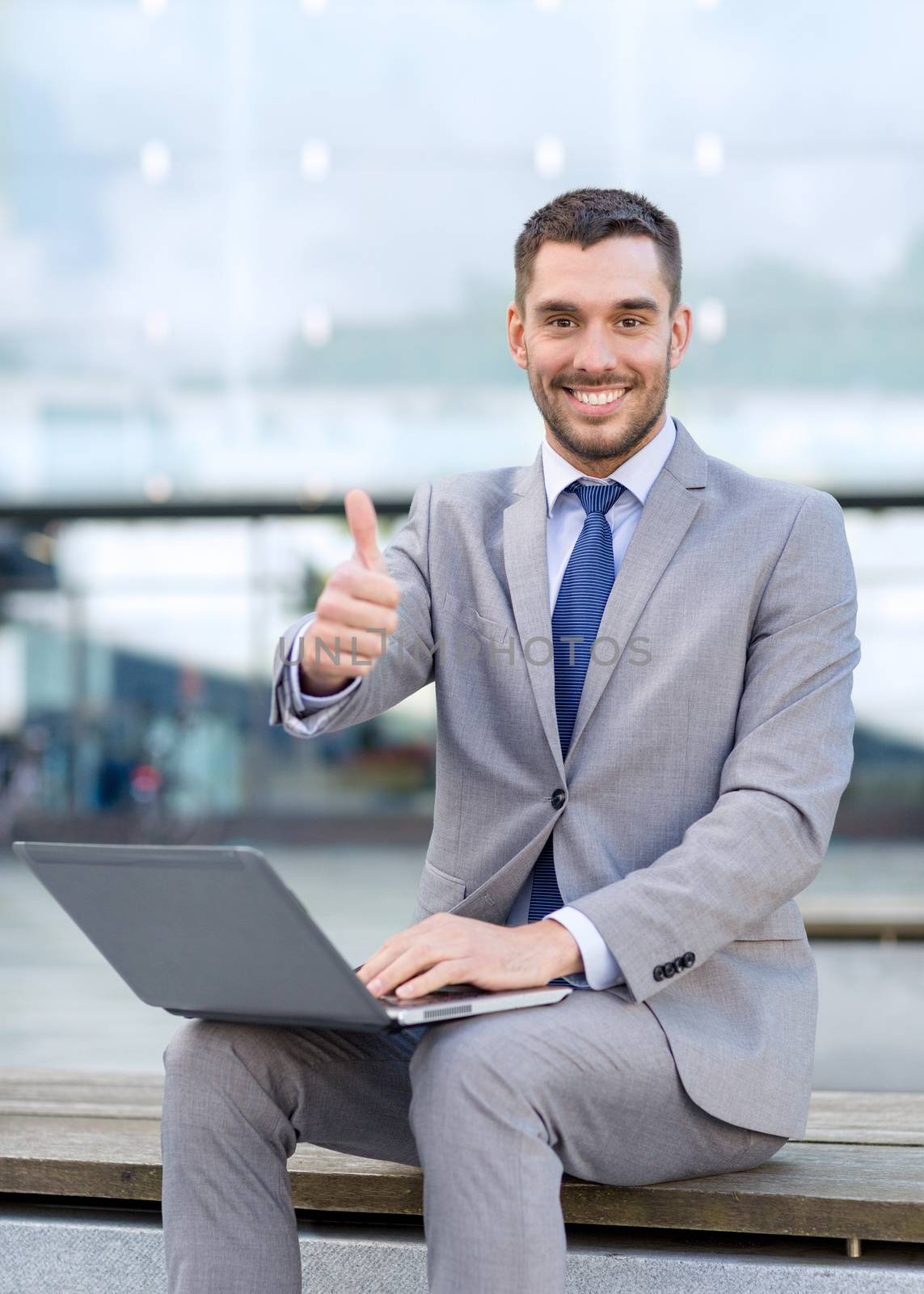 business, education, technology, gesture and people concept - smiling businessman working with laptop computer showing thumbs up on city street