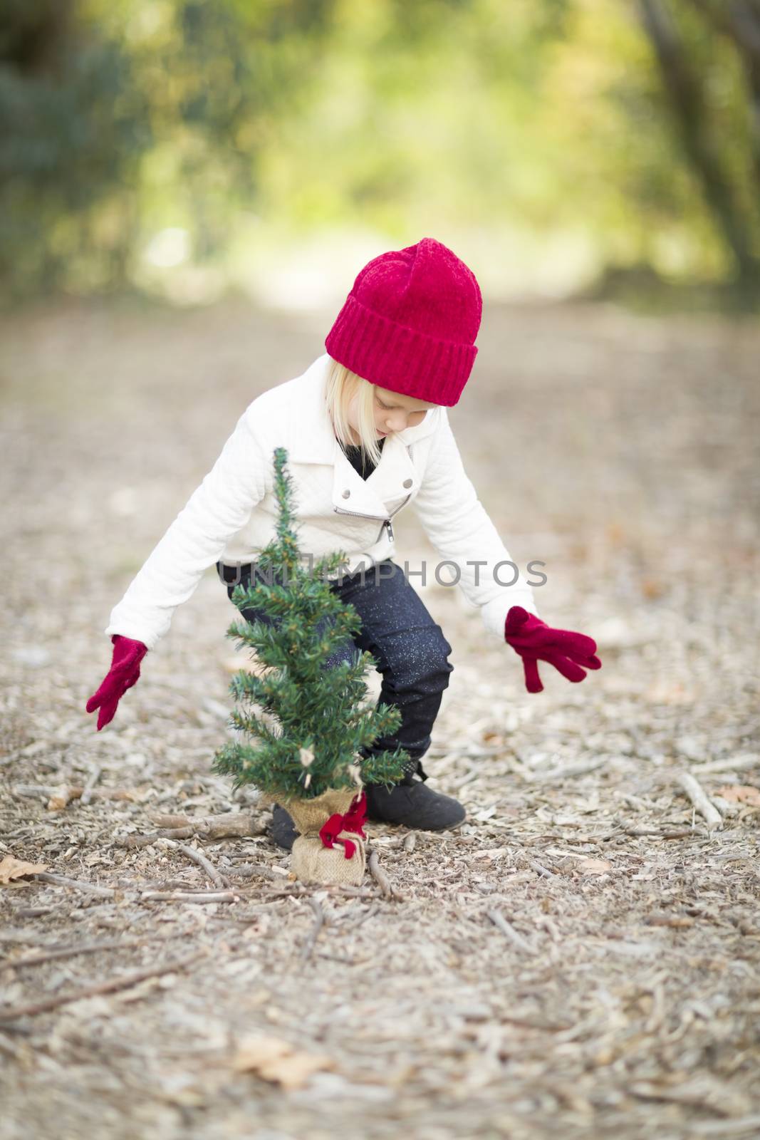 Baby Girl In Red Mittens and Cap Near Small Christmas Tree Outdoors.
