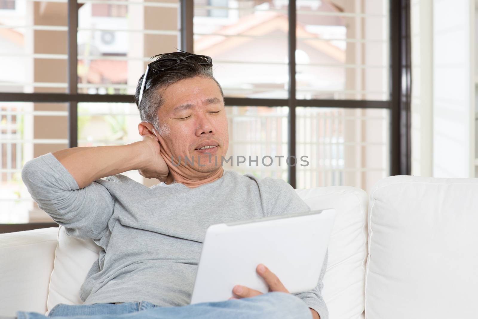 Portrait of 50s mature Asian man shoulder pain, pressing on neck with tired expression after long period using tablet computer, sitting on sofa at home.