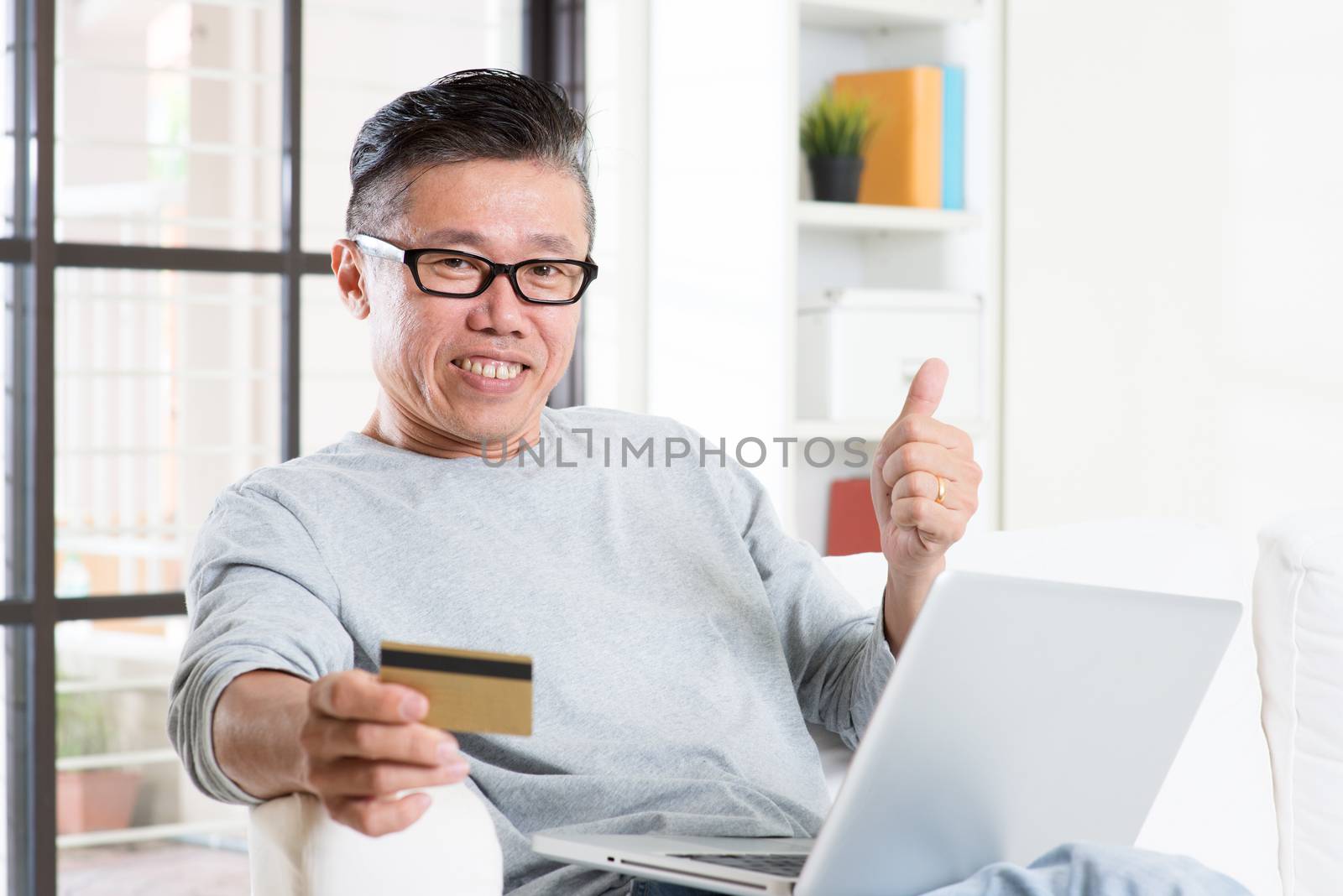 Portrait of 50s mature Asian man using computer doing online payment with credit card and thumb up, sitting on sofa at home, senior retiree indoors living lifestyle.
