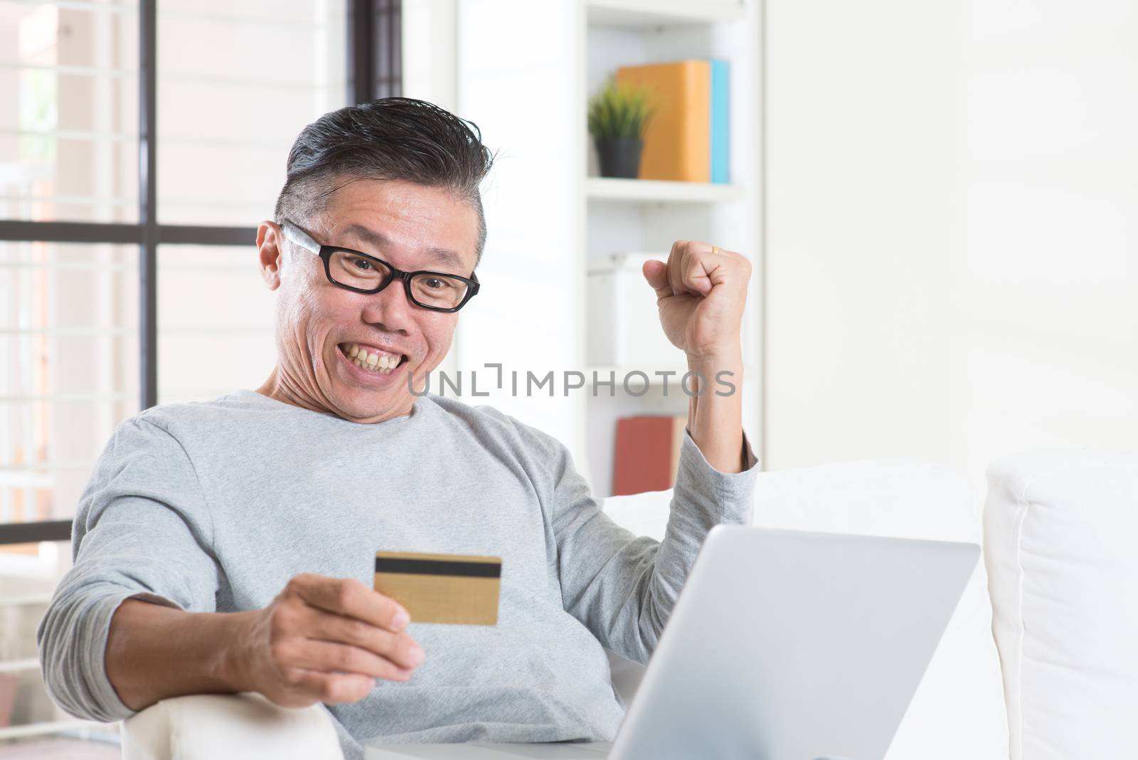 Portrait of 50s mature Asian man winning prizes while using computer internet doing online payment with credit card, sitting on sofa at home.