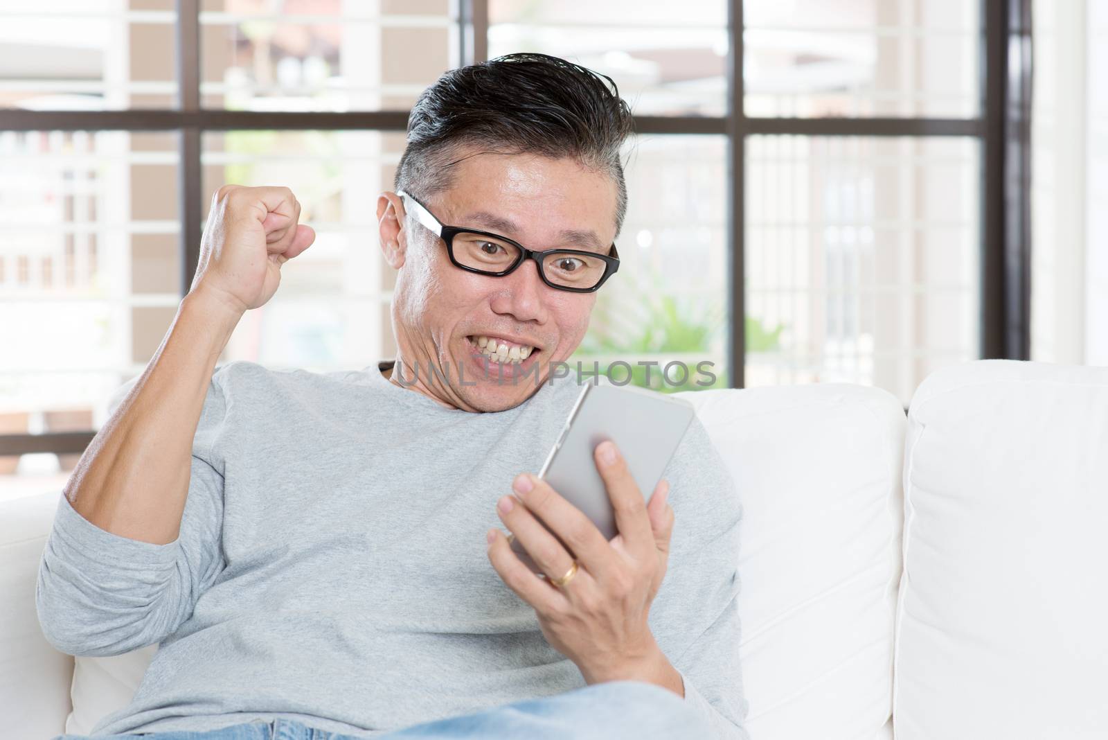 Portrait of excited 50s mature Asian man celebrating success while using smart phone, sitting on sofa at home.