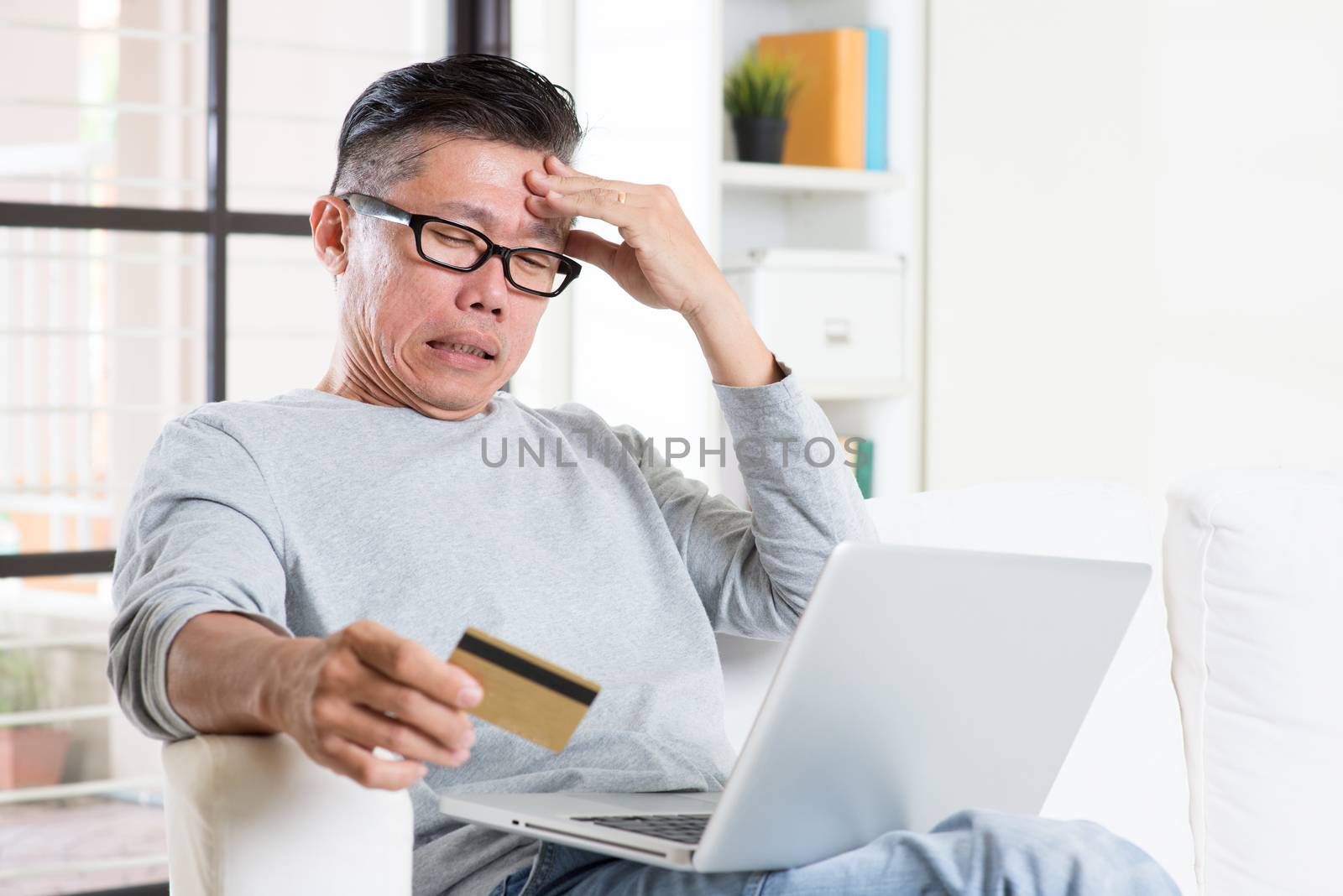 Portrait of 50s mature Asian man having problems while using computer internet doing online payment with credit card, sitting on sofa at home.