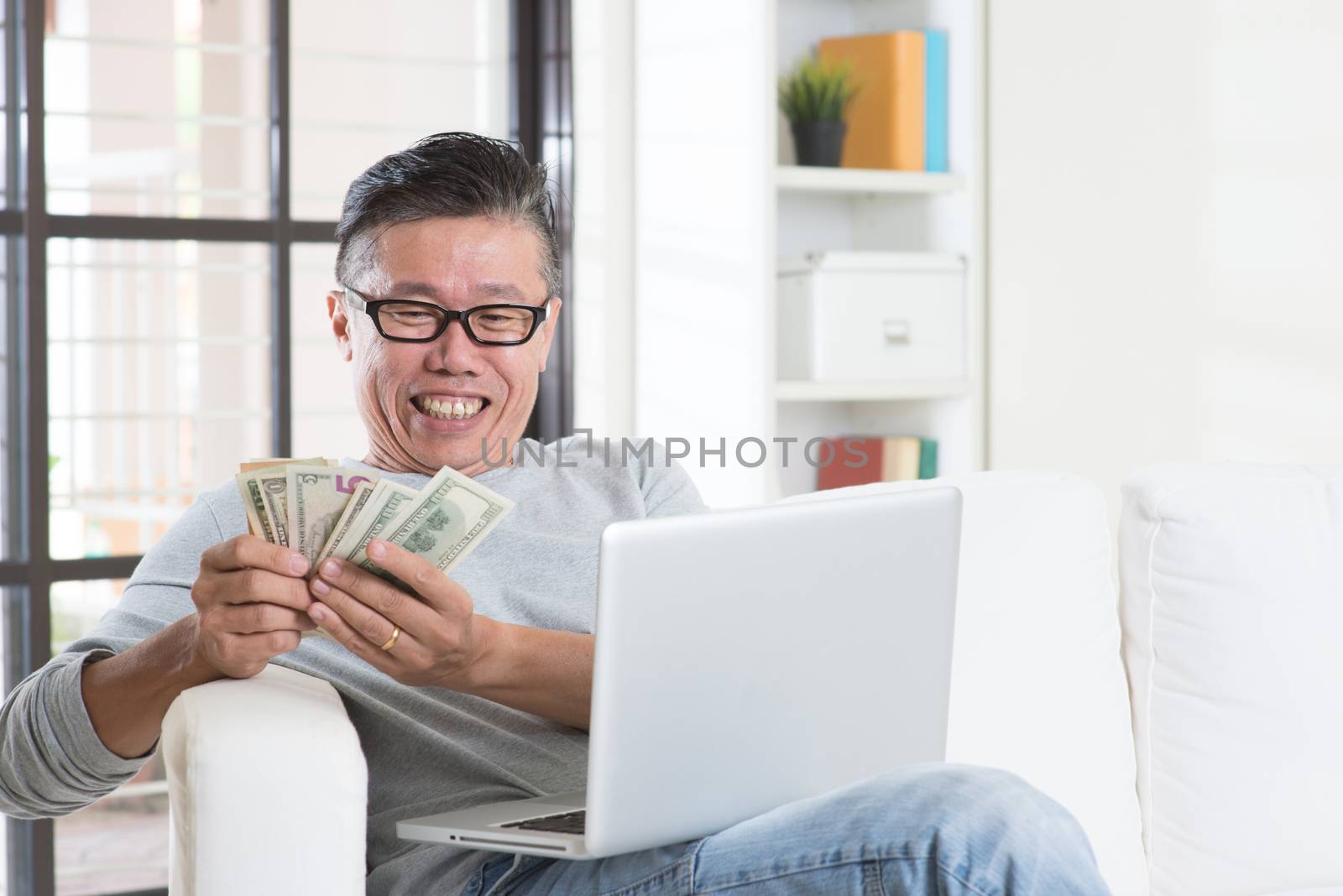 Portrait of happy 50s mature Asian man using internet computer and counting cash at home, earning money from his successful online business. Working from home concept.