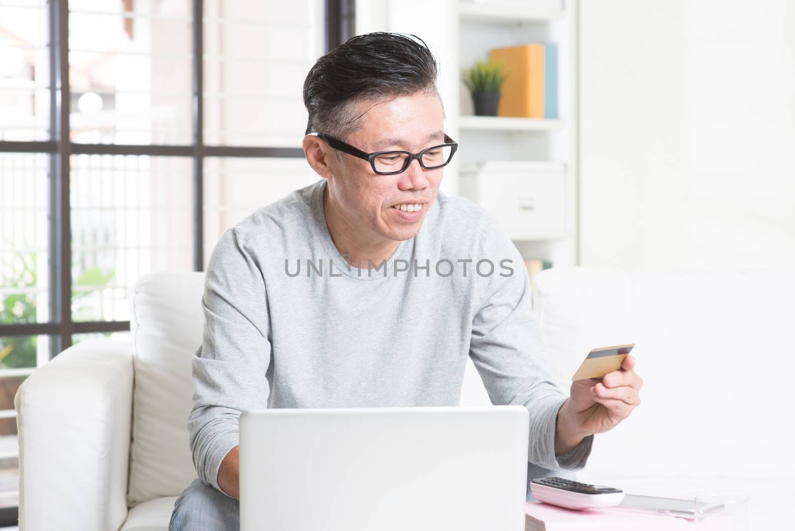 Portrait of 50s mature Asian man using computer doing online shopping with credit card, sitting on sofa at home, senior retiree indoors living lifestyle.