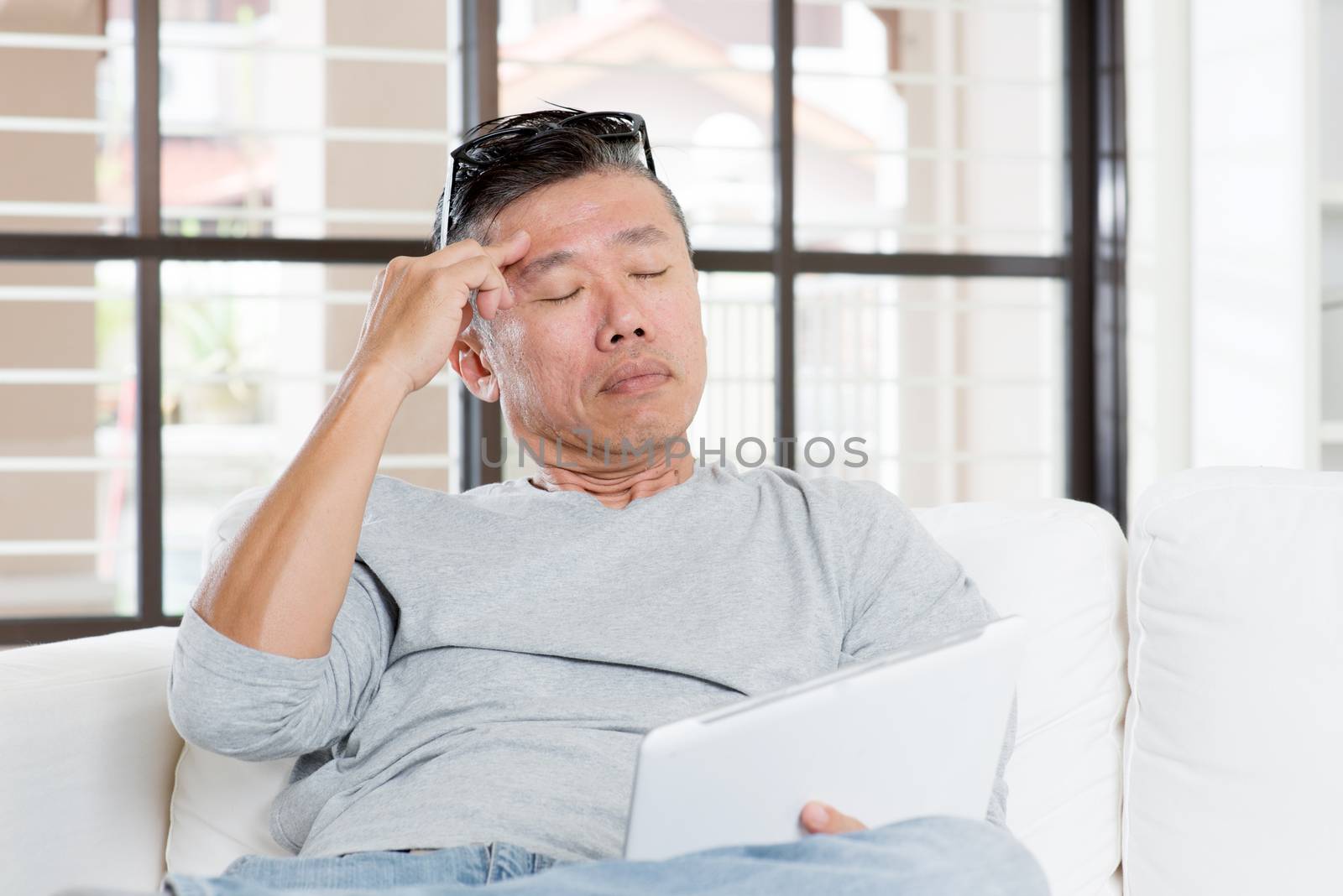 Portrait of 50s mature Asian man headache, pressing on head with tired expression after long period using tablet computer, sitting on sofa at home.