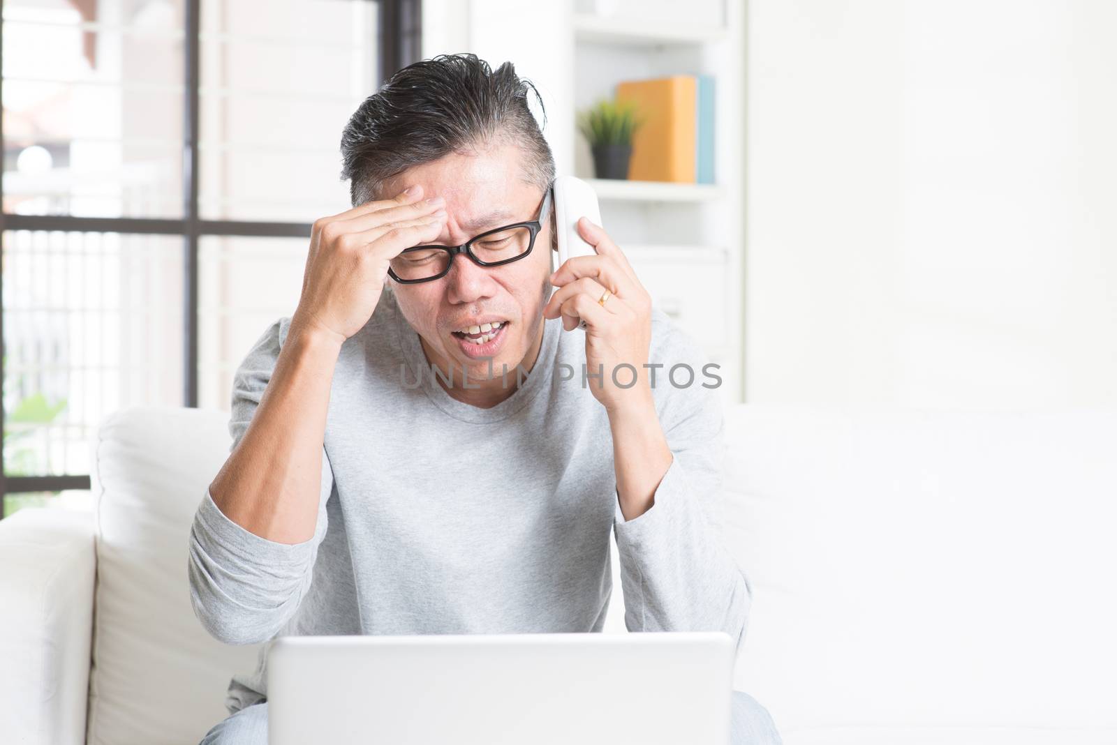 Portrait of 50s mature Asian man having problems and calling on phone, using computer internet working from home, sitting on sofa indoors.