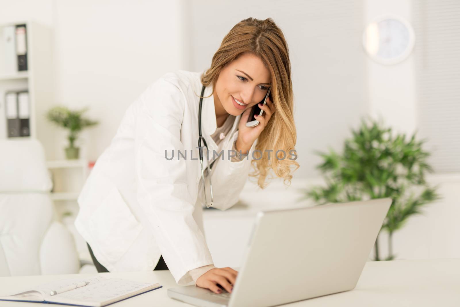 Beautiful young female doctor in her consulting room, using phone and writing on laptop.