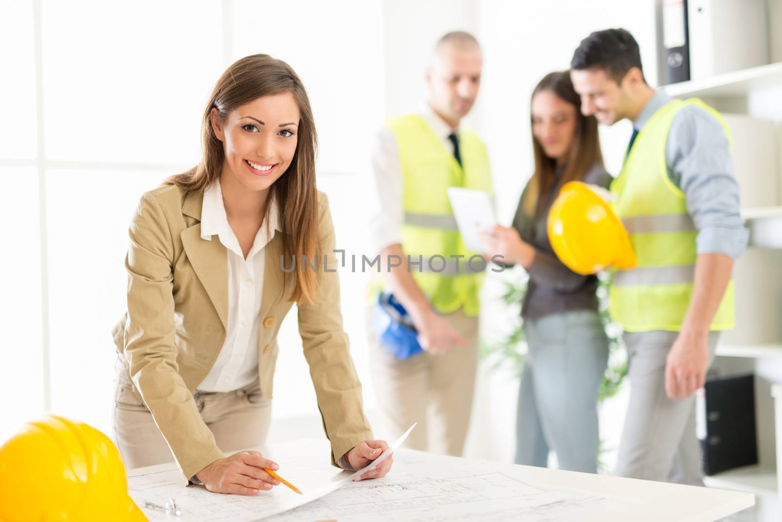 Smiling female architect analyzing blueprint at desk in office with colleagues in background. Looking at camera.