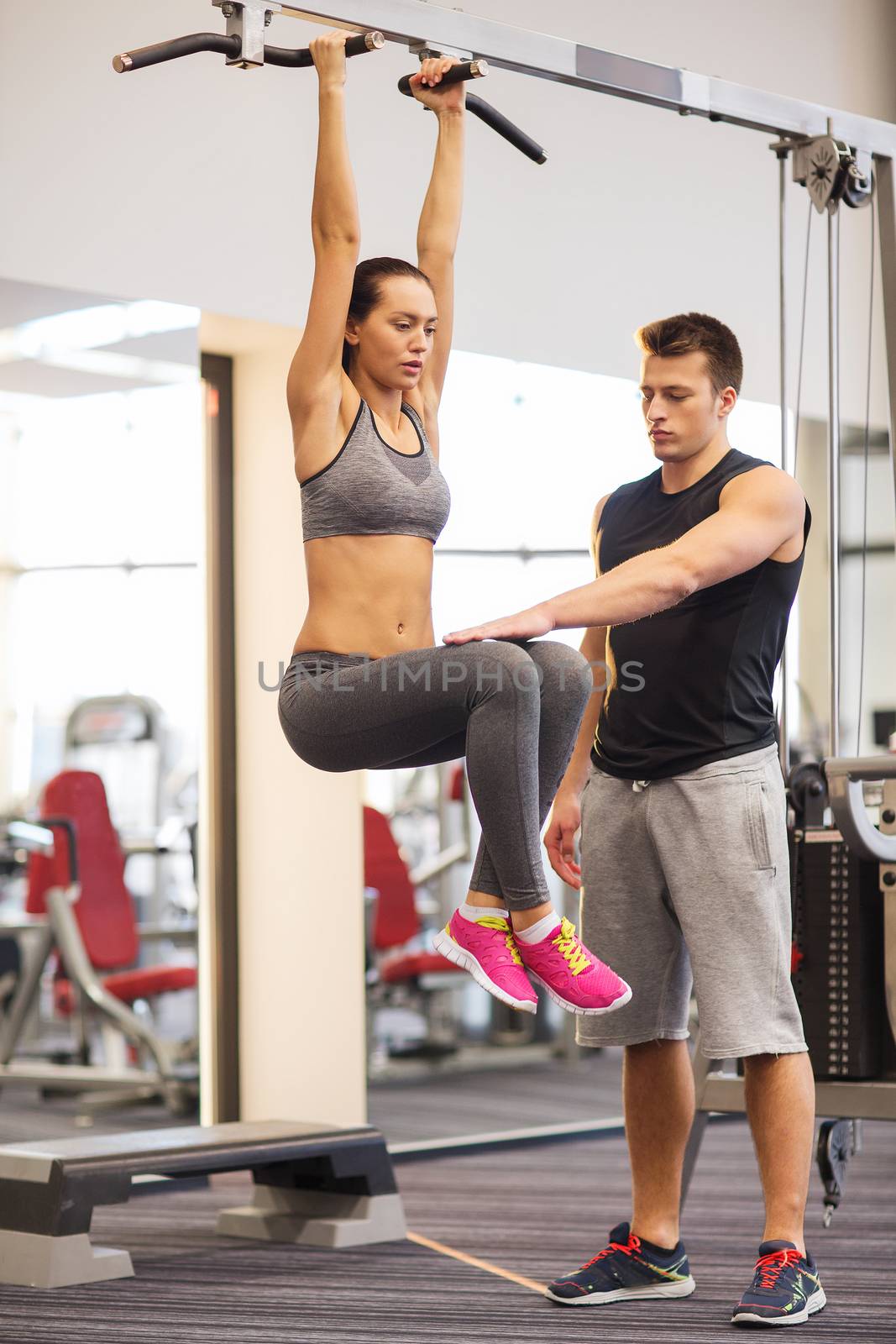 sport, fitness, lifestyle, teamwork and people concept - young woman with trainer hanging on bar and doing leg raises in gym