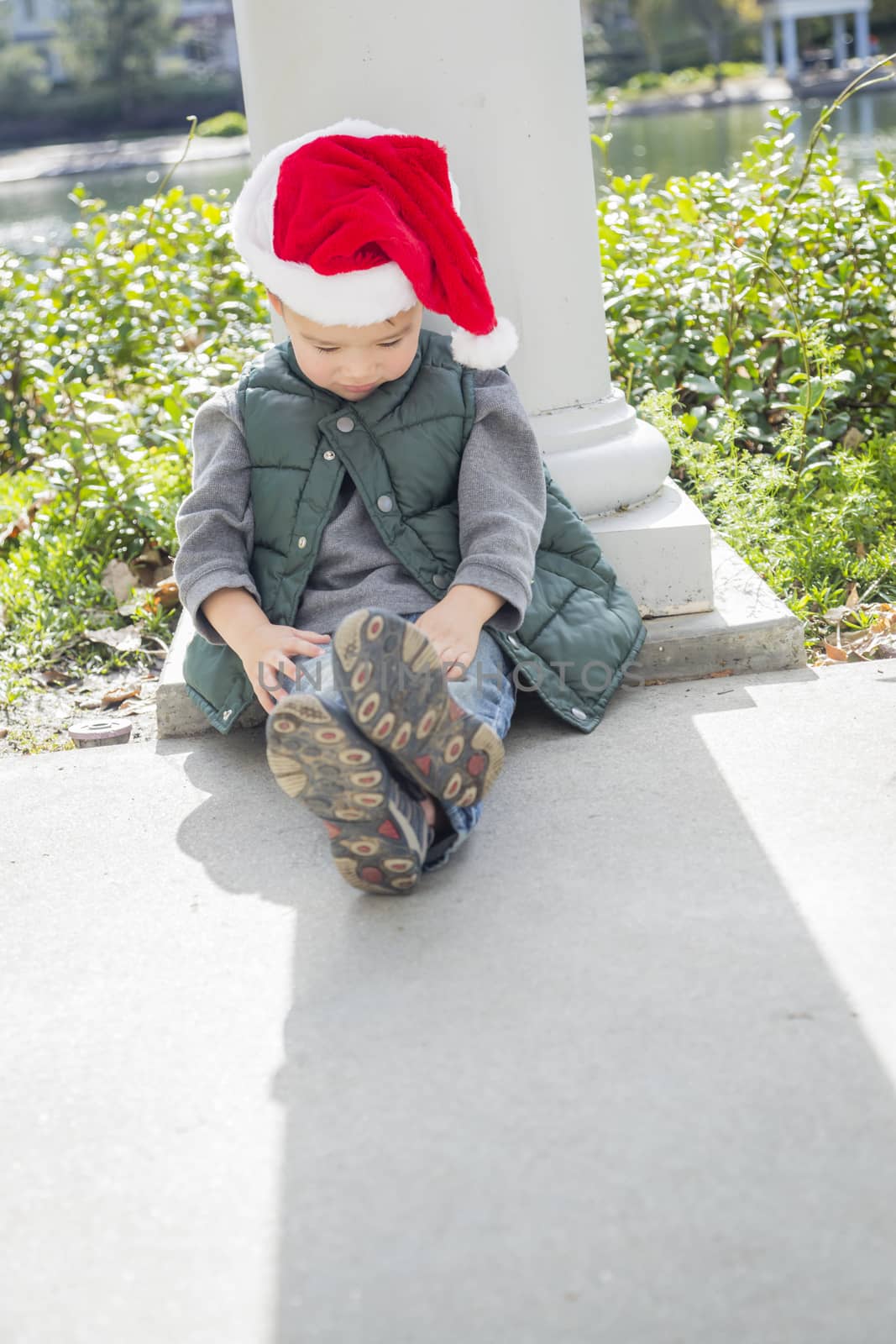 Melancholy Mixed Race Boy Sitting At The Park Wearing Christmas Santa Hat.
