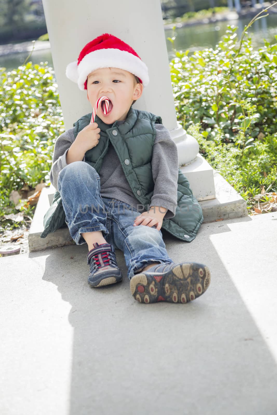 Cute Mixed Race Boy Sitting Wearing Christmas Santa Hat and Enjoying A Candy Cane.