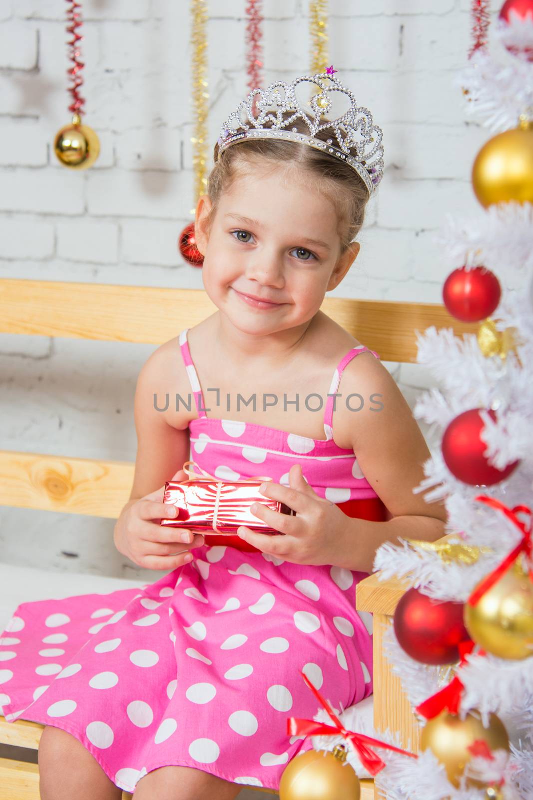 Four-year girl sitting on a bench with a small gift from snowy Christmas trees