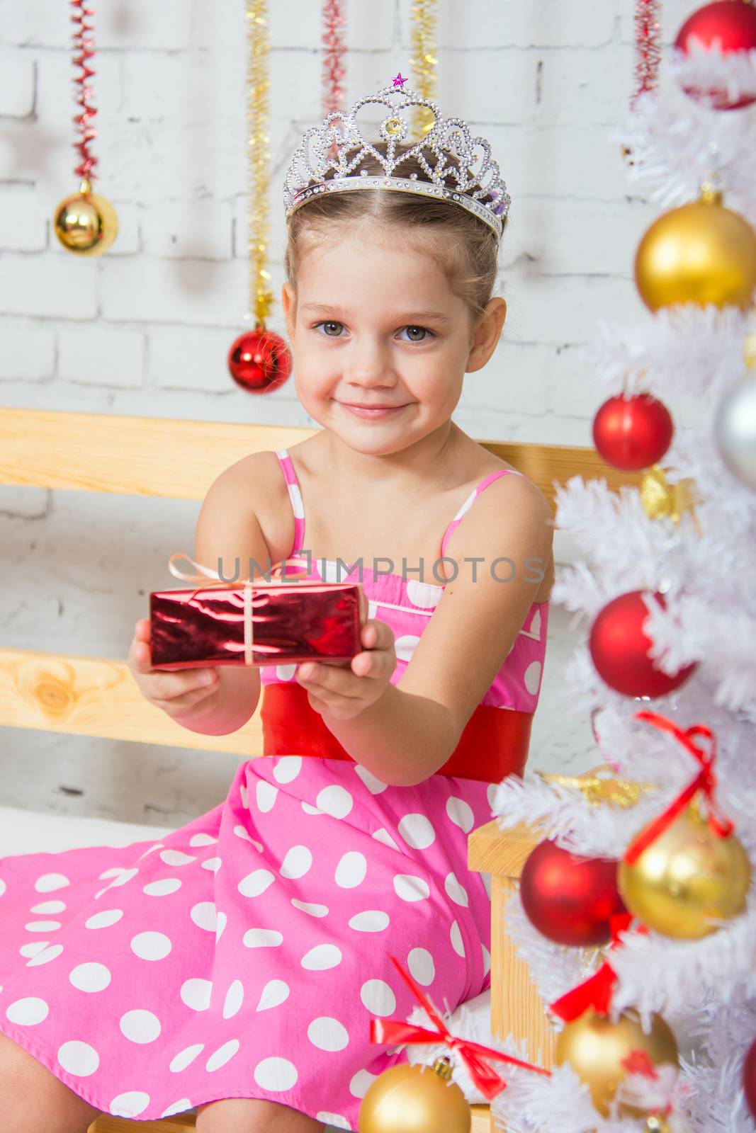 Four-year girl sitting on a bench with a small gift from snowy Christmas trees