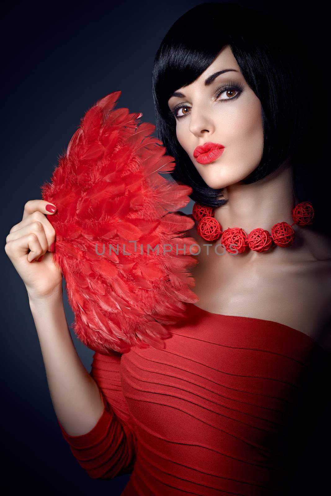 Fashion beauty portrait of sexy brunette woman, silky bob hairstyle on dark. Sensual lady with stylish necklace red feather fan sending you a kiss, makeup, lips. Vivid people                          